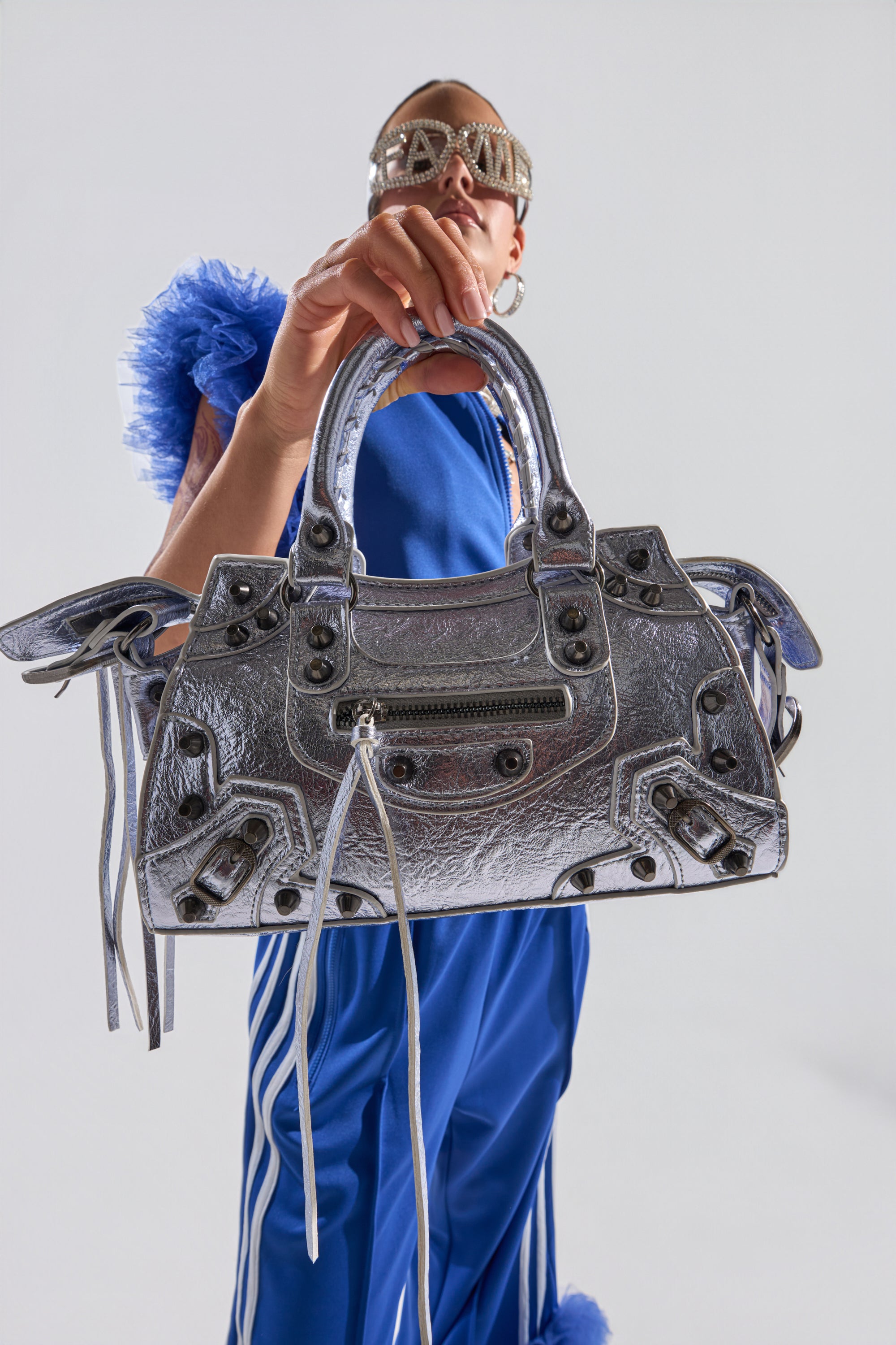 Wearing a blue outfit and statement sunglasses, a person presents the TO THE CITY PURSE—a large metallic faux leather bag with a removable shoulder strap—toward the camera against a plain white background. 1