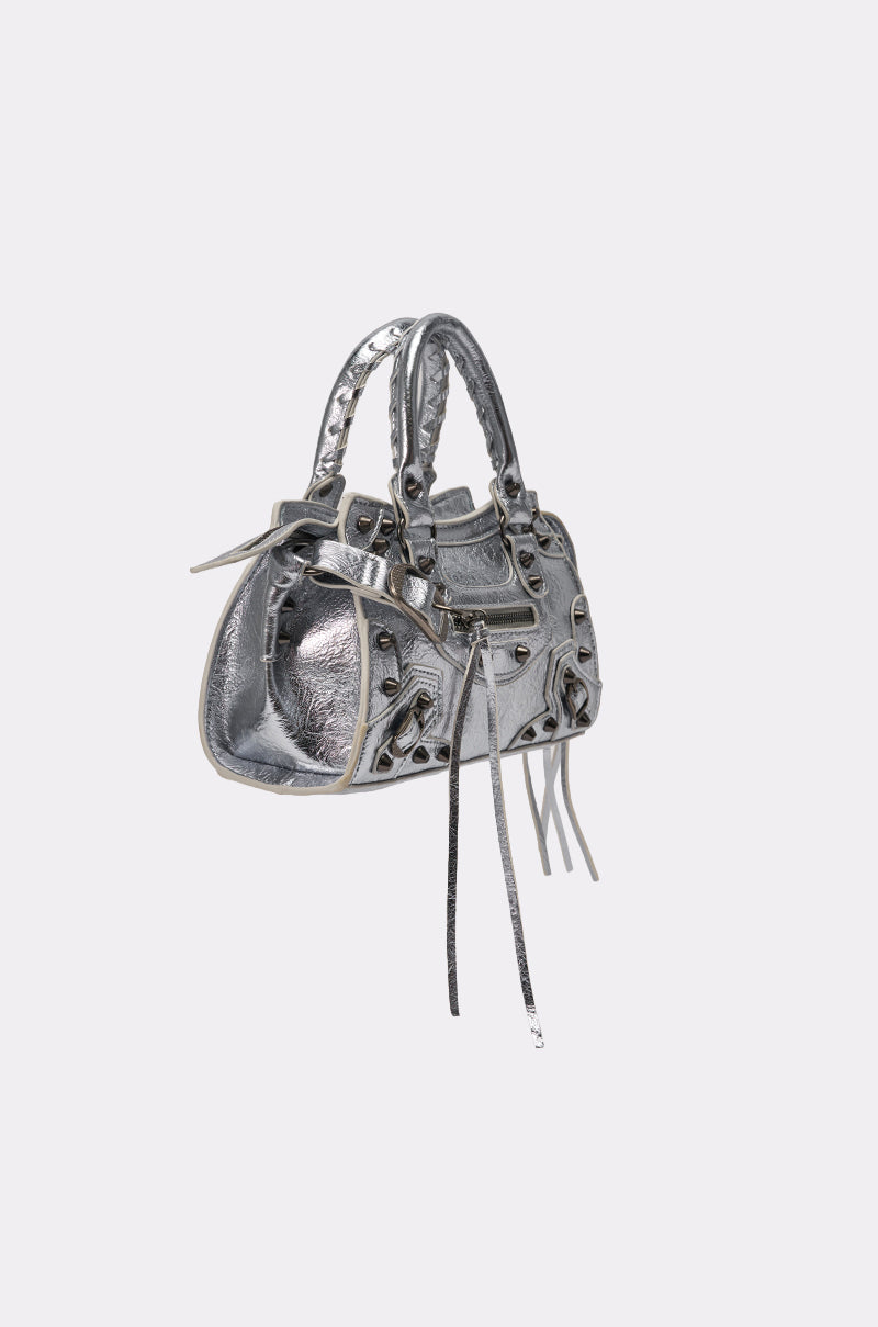 The TO THE CITY PURSE is a small metallic faux leather bag in silver with braided handles, decorative buckles, zipper accents, long tassels, and a removable shoulder strap, shown on a plain light gray background. 4