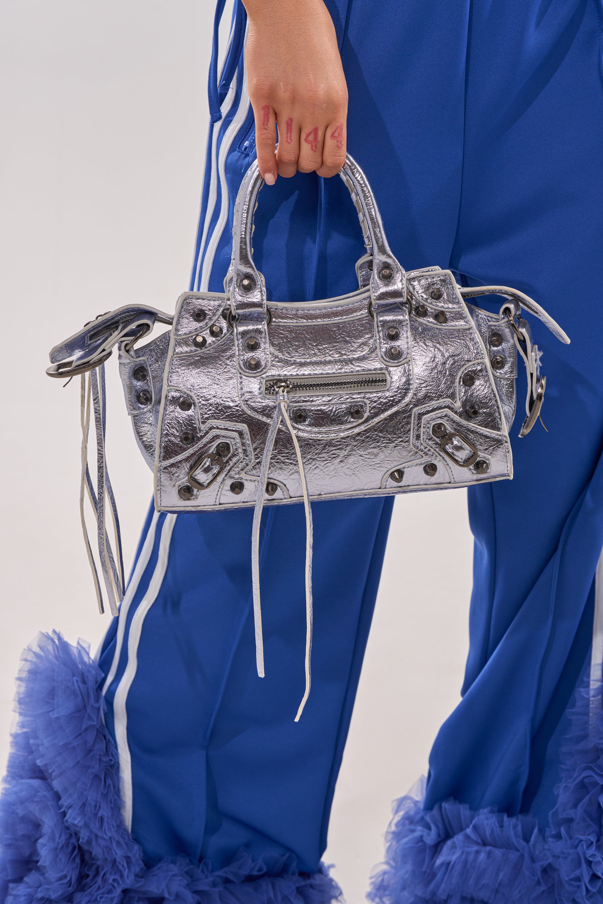 A person in blue pants with white stripes and ruffled hems holds the TO THE CITY PURSE—a faux leather metallic bag featuring studs, zipper details, long leather pulls, and a removable shoulder strap. 2