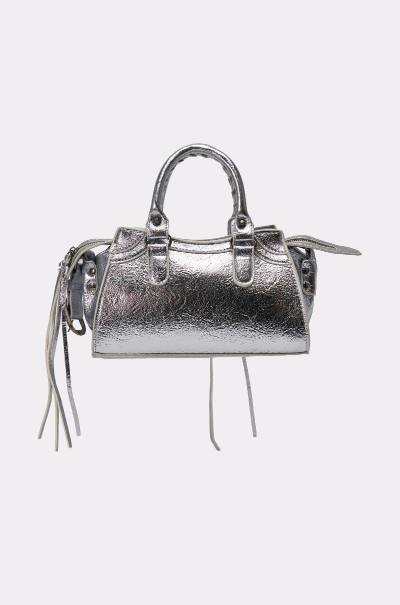 The TO THE CITY PURSE is a small silver faux leather bag with two short handles, side zipper accents, decorative tassels, and a removable shoulder strap, shown against a plain light background. 5