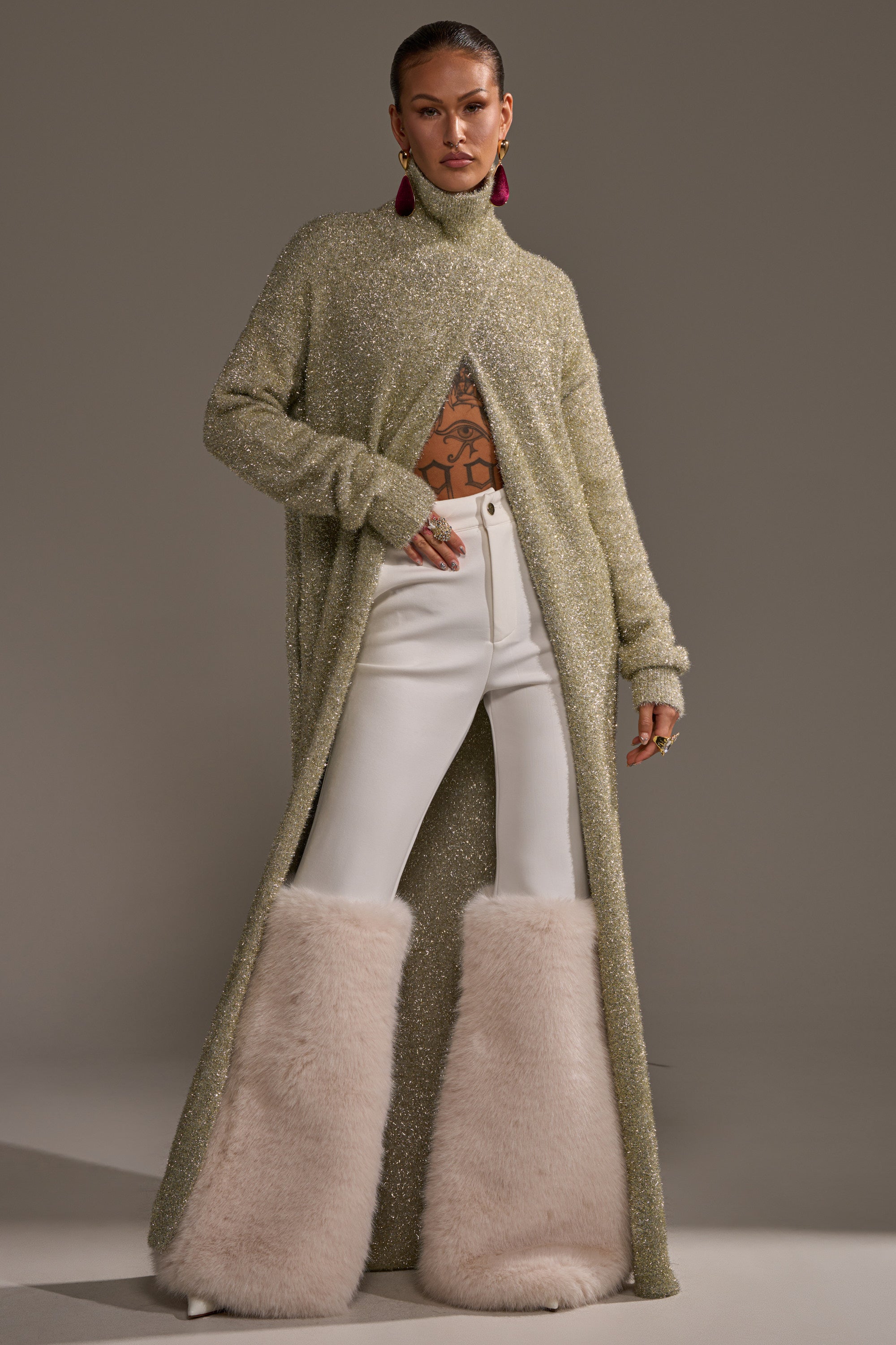 A model wears the WORTHY OF LOVE LONG SLEEVE METALLIC SWEATER IN GOLD under a sparkly open-front coat and white high-waisted pants with pale fur at the lower legs, accessorized with bold earrings and showcasing visible torso tattoos. 1