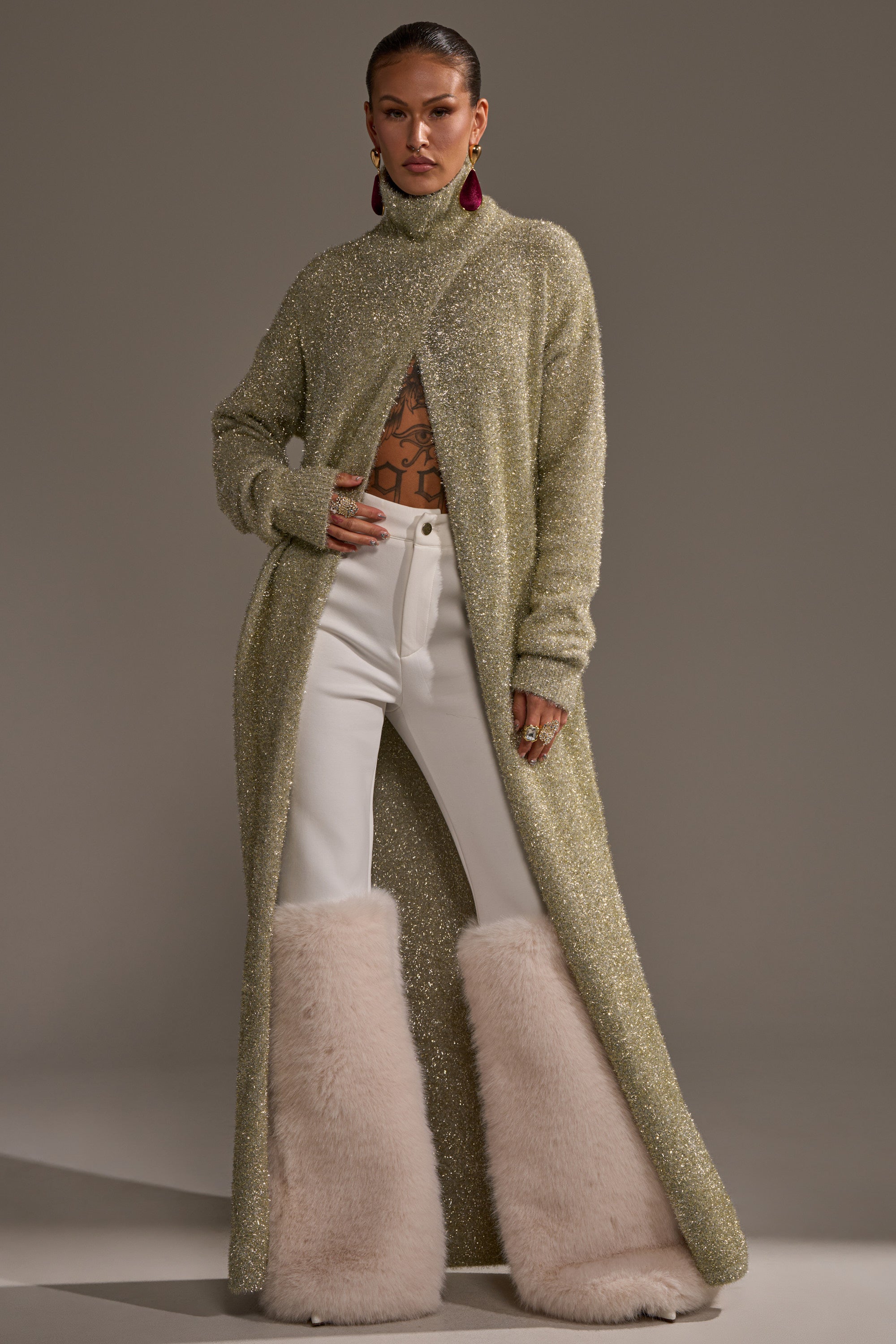 A woman poses in the WORTHY OF LOVE LONG SLEEVE METALLIC SWEATER IN GOLD over a bare chest with tattoos, paired with white high-waisted pants and tall, fluffy cream boots against a plain background. 2