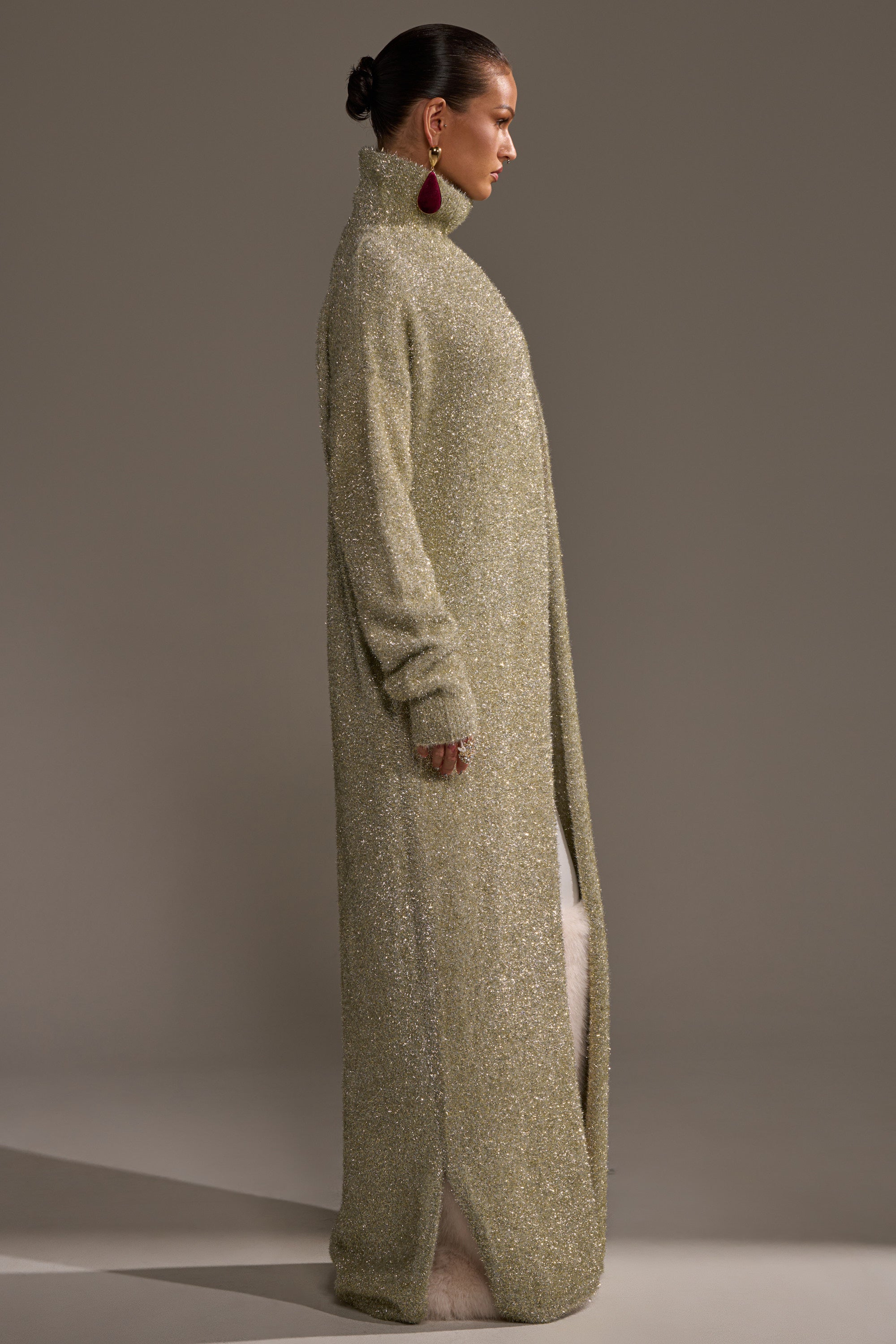 A woman in profile wears the WORTHY OF LOVE LONG SLEEVE METALLIC SWEATER IN GOLD, styled with her hair in a bun and statement earrings, set against a simple neutral background. 3