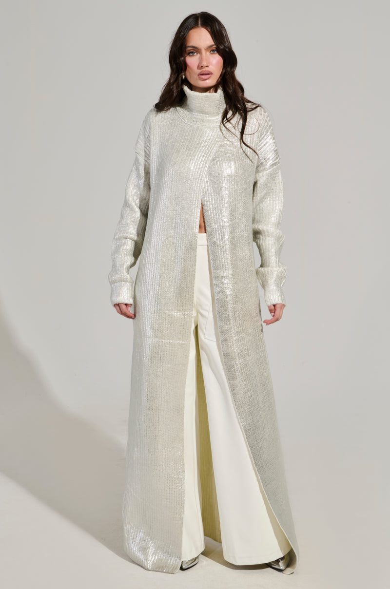 A person stands against a plain backdrop, wearing the Worthy of Love Long Sleeve Metallic Sweater in Silver under a long, shiny silver coat and paired with wide-leg white pants.