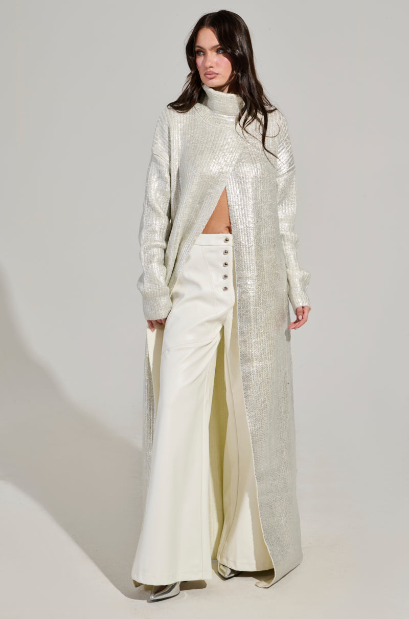 A person wearing a shimmering long silver coat with a high collar and front slit, paired with high-waisted, wide-leg white pants adorned with buttons. The ensemble is complemented by the "Worthy of Love Long Sleeve Metallic Sweater in Silver," standing against a plain background.