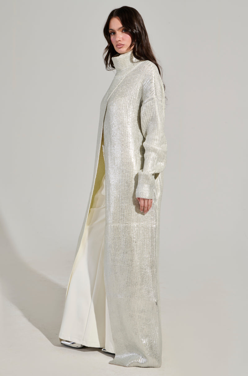 A person with long brown hair is wearing the "Worthy of Love" long sleeve metallic sweater in silver, paired with white pants. They are standing against a plain light background.