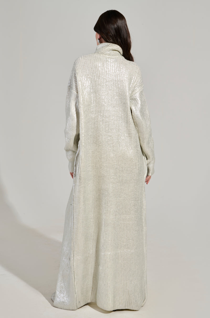 A person stands with their back turned, dressed in the WORTHY OF LOVE LONG SLEEVE METALLIC SWEATER IN SILVER. This shimmering silver sweater coat, resembling a turtleneck maxi design, features a high collar and extends gracefully to the floor, showcasing a slightly textured finish. The person's long dark hair provides a striking contrast to the shiny fabric.