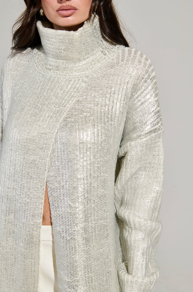 The photo showcases a person in a WORTHY OF LOVE LONG SLEEVE METALLIC SWEATER IN SILVER, featuring textured knit details and a turtleneck design. The image highlights the upper body under soft lighting, emphasizing the light-colored maxi sweater's partial open front.