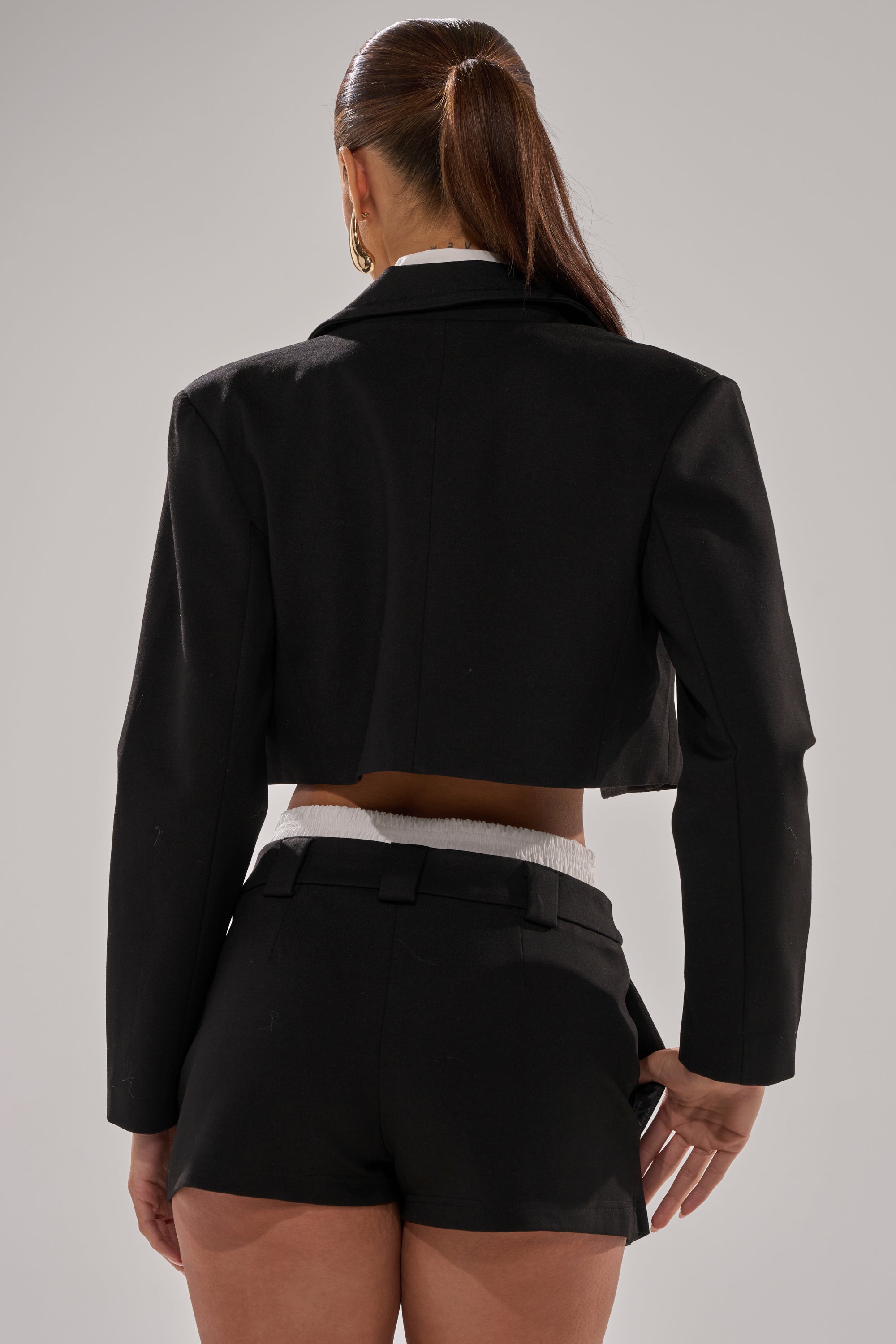 A woman with long brown hair in a ponytail is shown from the back, wearing the BLACK TIE FORMAL TOP and high-waisted black shorts against a neutral background.
