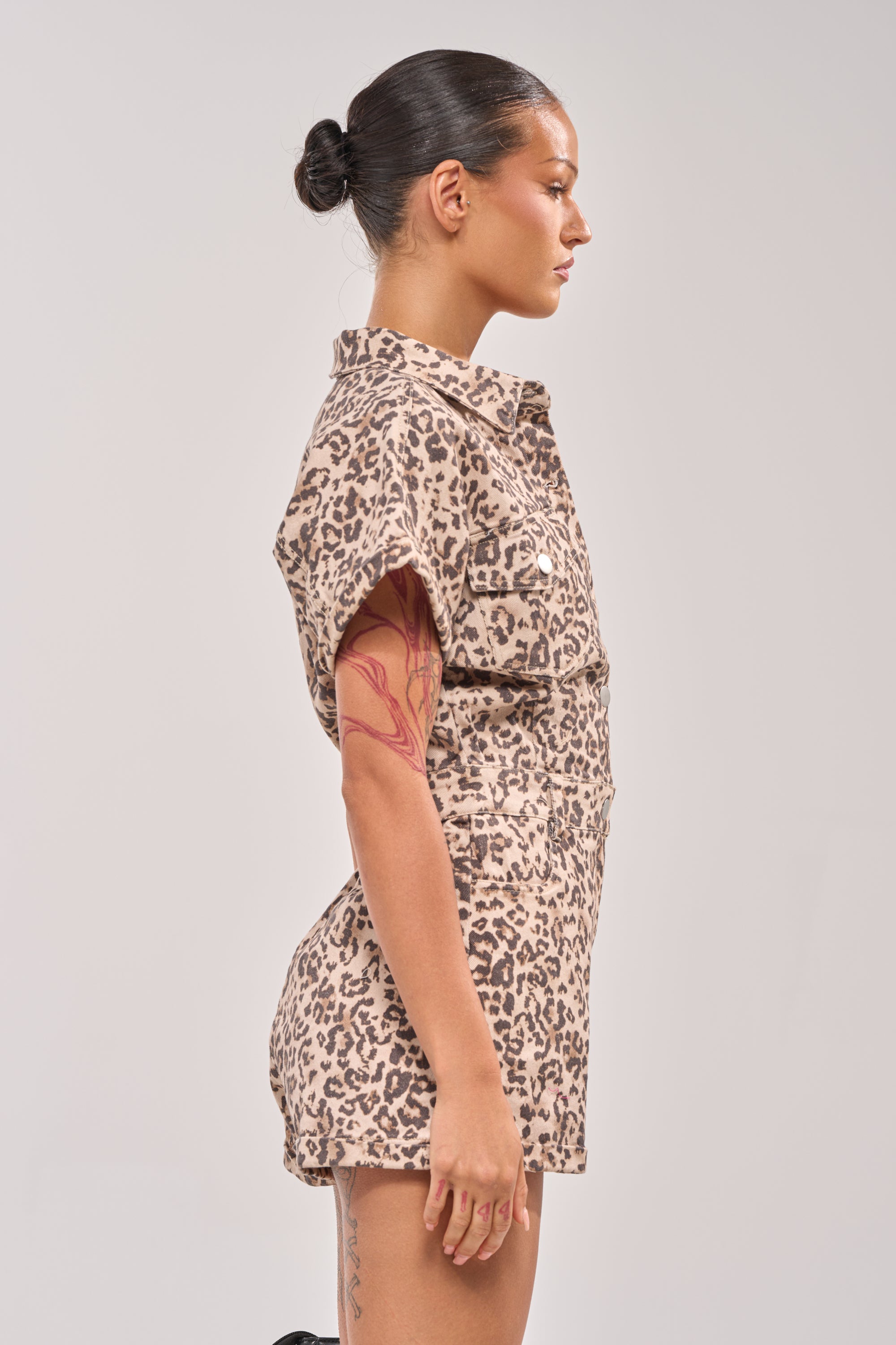 A woman with dark hair in a neat bun models the SHADES ON DENIM ROMPER IN LEOPARD, featuring short sleeves and a bold print. She displays tattoos on her left arm and thigh while posing against a plain light backdrop. 2
