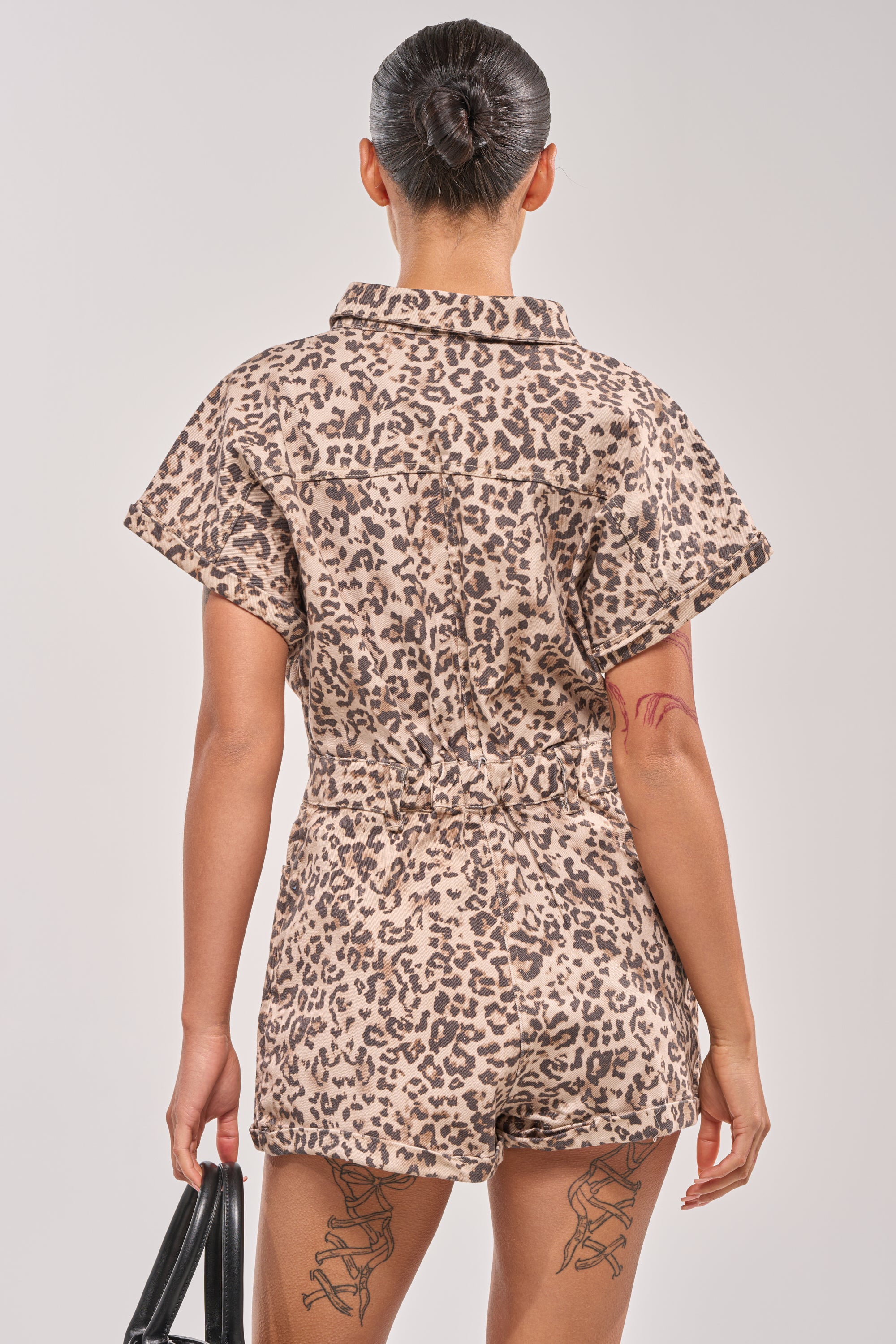 A person with their hair in a bun is seen from the back, holding a black handbag and showing off tattoos while wearing the SHADES ON DENIM ROMPER IN LEOPARD, embodying chic utility style. 3