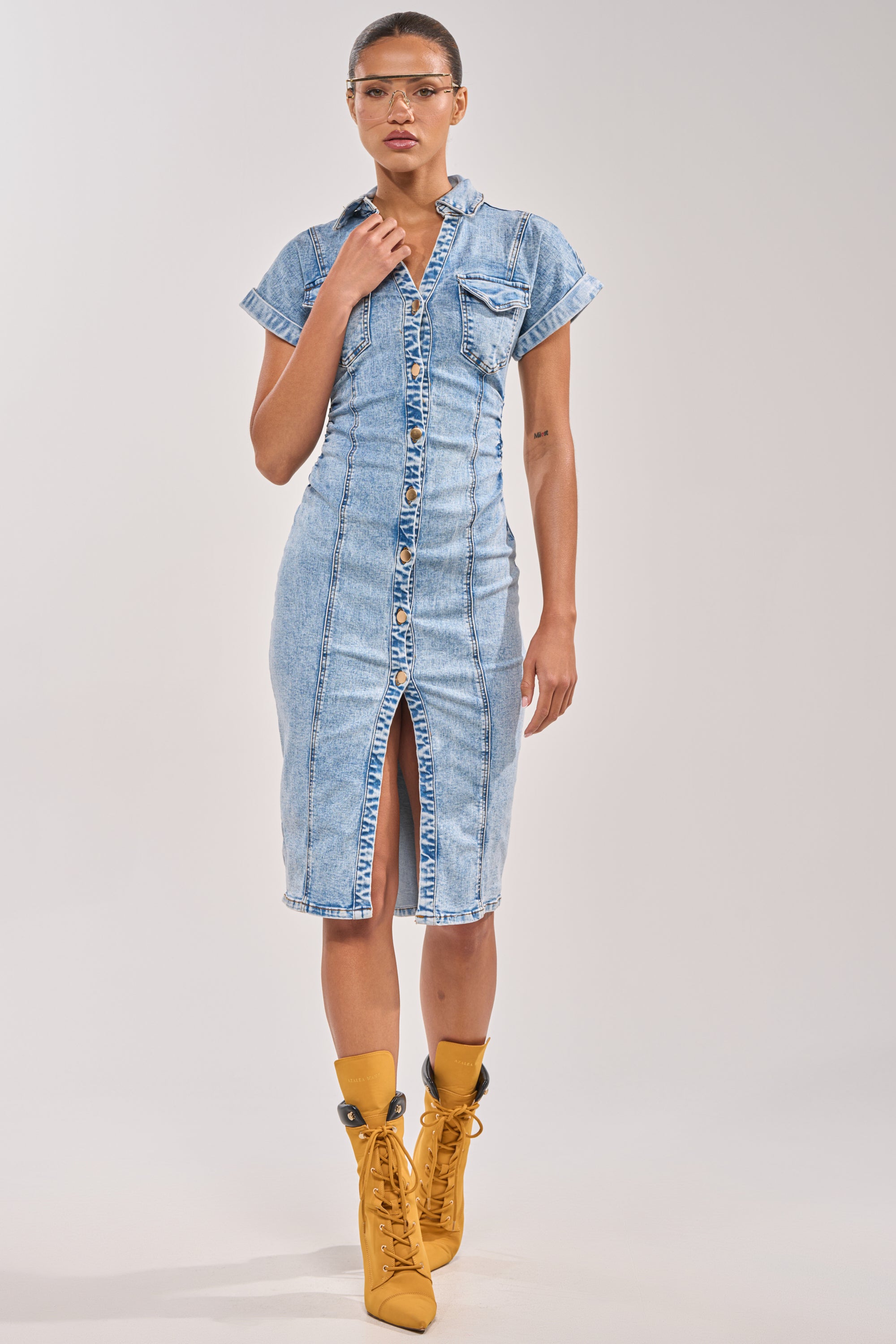 Wearing the LITTLE DENIM MIDI DRESS IN LIGHT BLUE with front pockets, a front slit, and curve-defining fit, a woman pairs it with yellow lace-up high-heeled boots and clear glasses against a plain background. 4