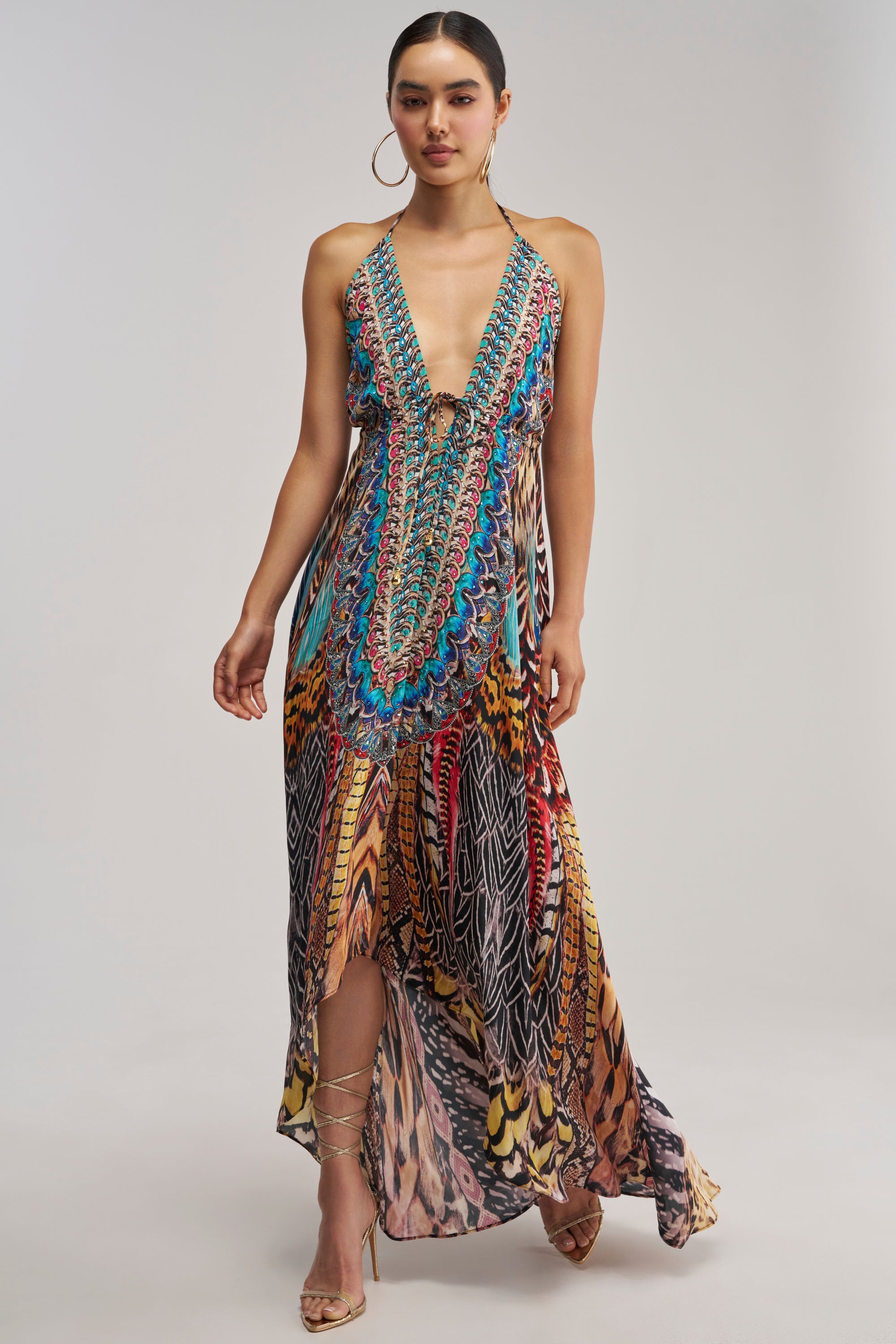 A woman wearing the TALK OF THE TOWN PRINTED MAXI DRESS, featuring a colorful pattern, deep V neckline, and high slit, paired with strappy heels, standing against a plain background.