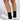 A person wears COZY CROSS SOCKS—black ribbed socks featuring a small white lizard design—with white cage-pattern sneakers, standing on a light background. Only the legs and feet are shown.