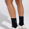 A person wears COZY CROSS SOCKS—black ribbed socks featuring a small white lizard design—with white cage-pattern sneakers, standing on a light background. Only the legs and feet are shown.