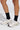 Someone stands on a light gray surface, showing only their lower legs and feet. They wear COZY CROSS SOCKS featuring metallic silver cross appliqué paired with chunky beige-and-white sneakers with cage-like overlays.