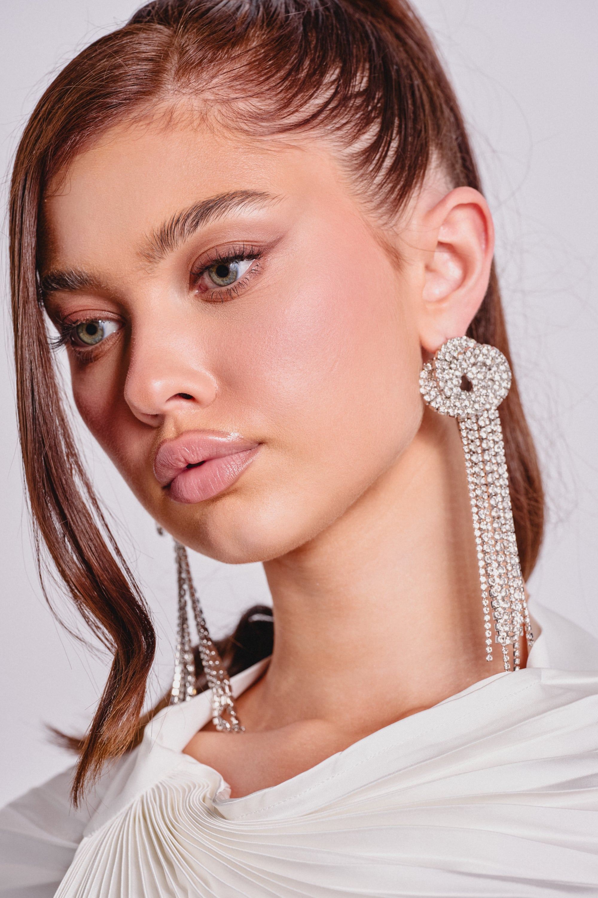 A woman with long brown hair in a high ponytail wears the AT THE PARTY EARRING—dramatic rhinestone fringe earrings—paired with a white pleated top and soft, neutral makeup for an elegant evening look as she gazes slightly downward. 0