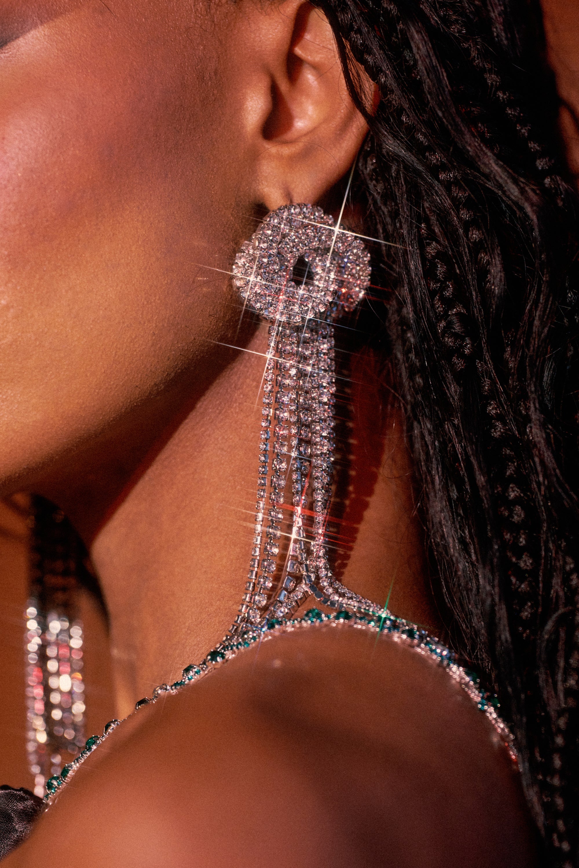 Close-up of a person with braided hair and glowing skin wearing the AT THE PARTY EARRING—large, sparkling statement earrings featuring rhinestone-studded, dangling strands that catch the light in a starburst effect. 3