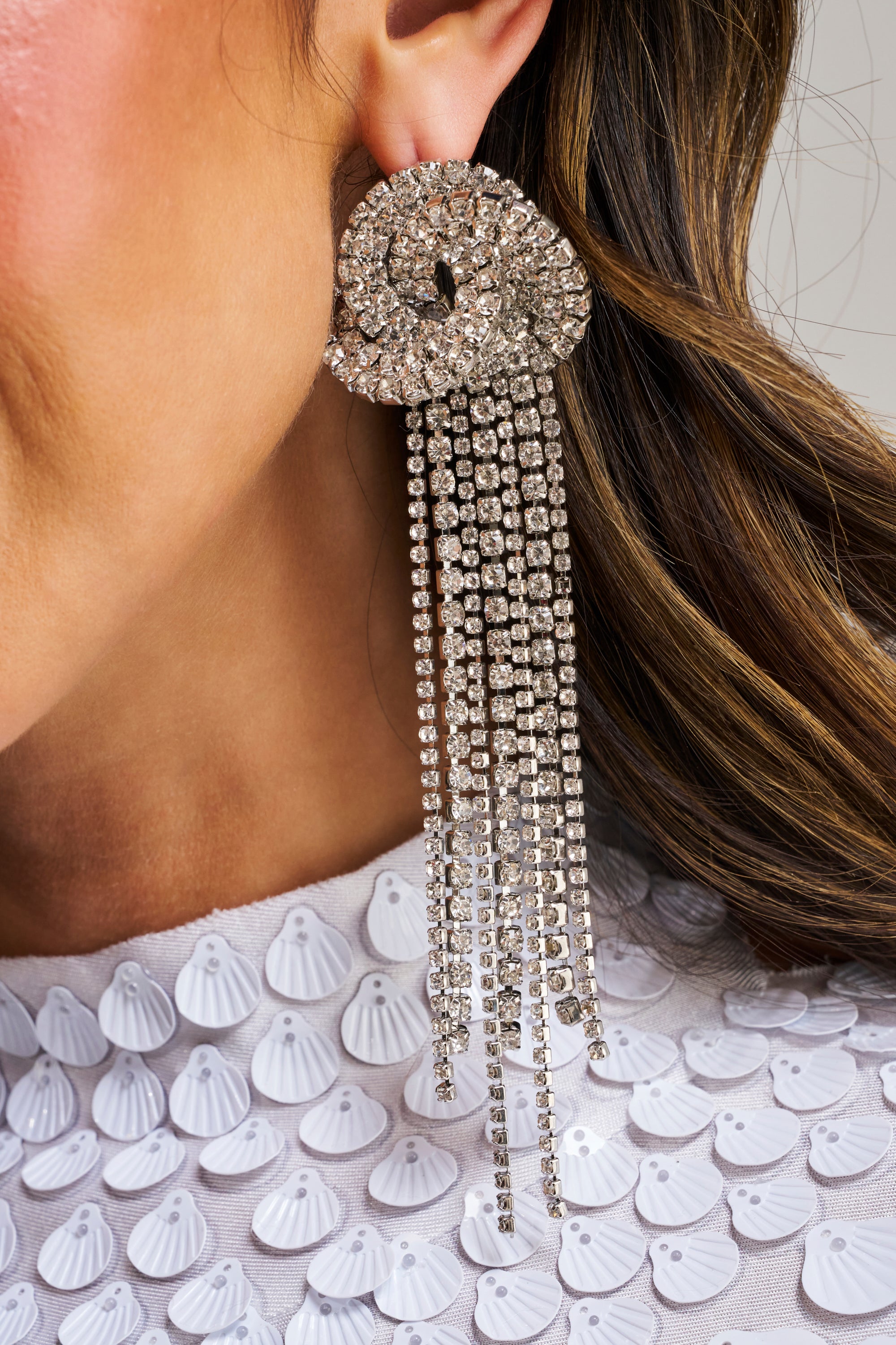 A close-up of a person wearing the AT THE PARTY EARRING, featuring statement rhinestone fringe with dangling strands. Their wavy brown hair and white top with shell-like details complete an elegant look ideal for evening occasions.