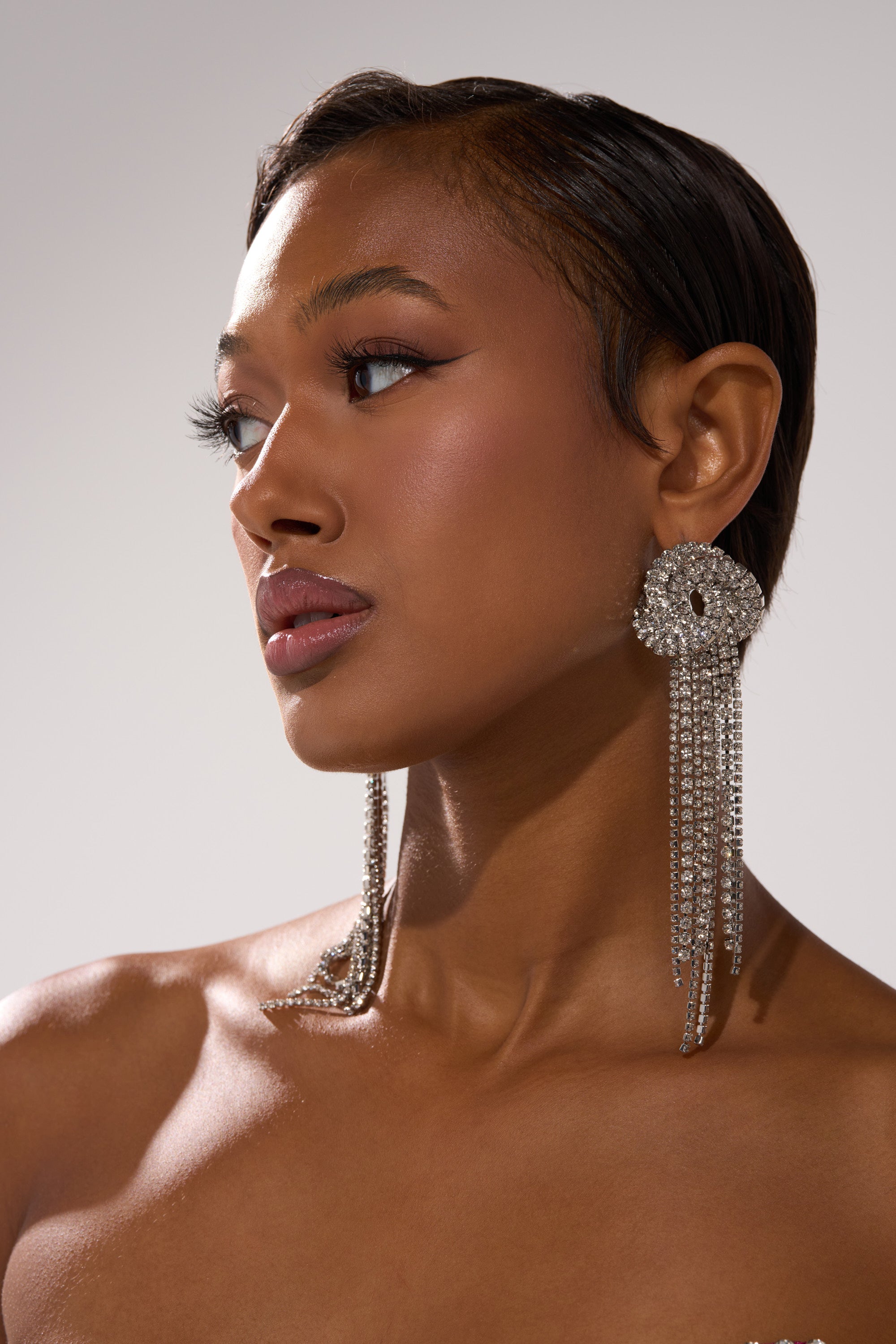 A woman with smooth, dark skin and short hair gazes to the side, wearing AT THE PARTY EARRING—dramatic fringe drop earrings with cascading chains—plus winged eyeliner and muted pink lipstick against a neutral background.