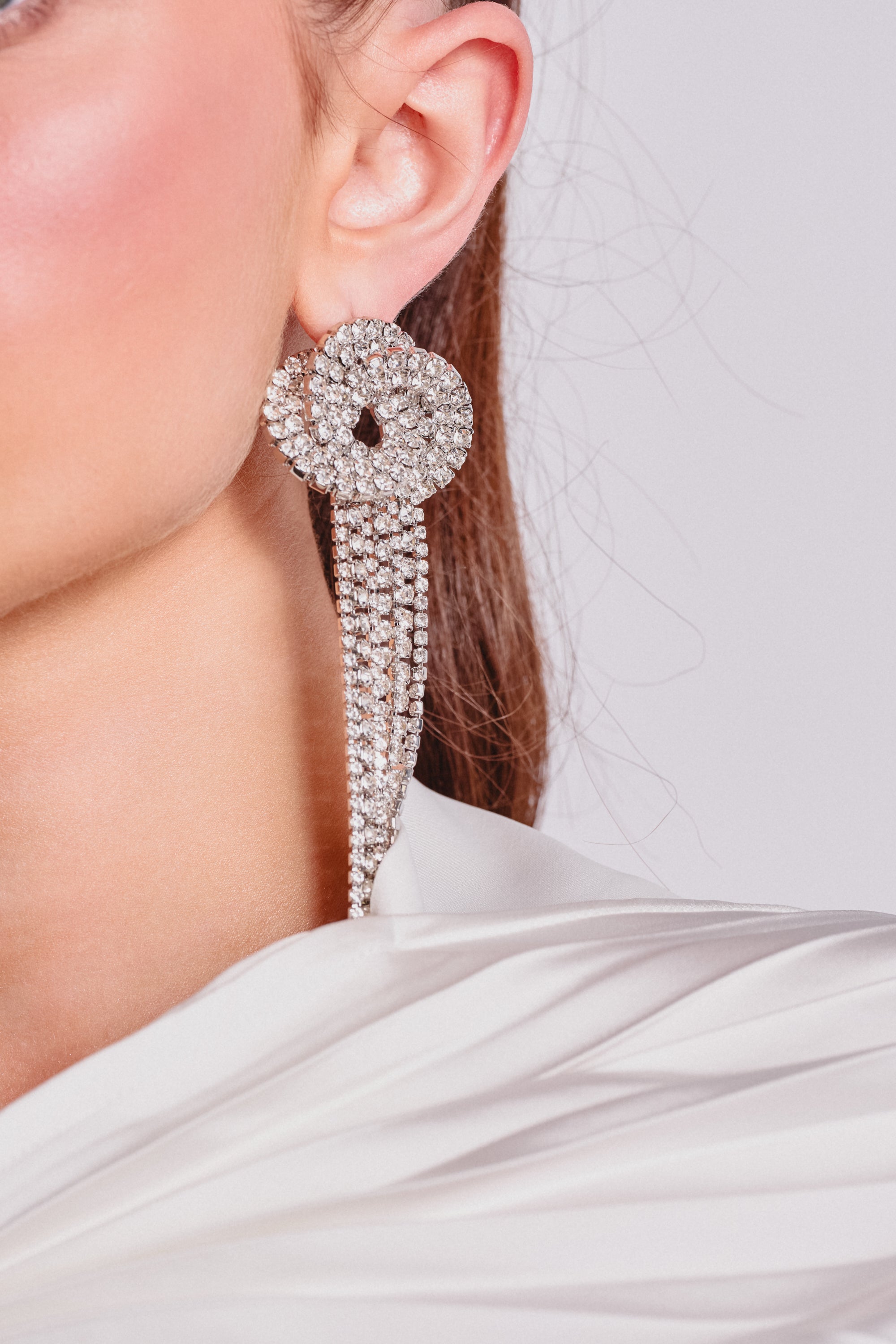 A close-up shows a woman's ear with the AT THE PARTY EARRING—rhinestone fringe earrings with long, dangling strands—paired with a white draped garment. Only part of her face and shoulder are visible. Perfect for enhancing evening looks. 1