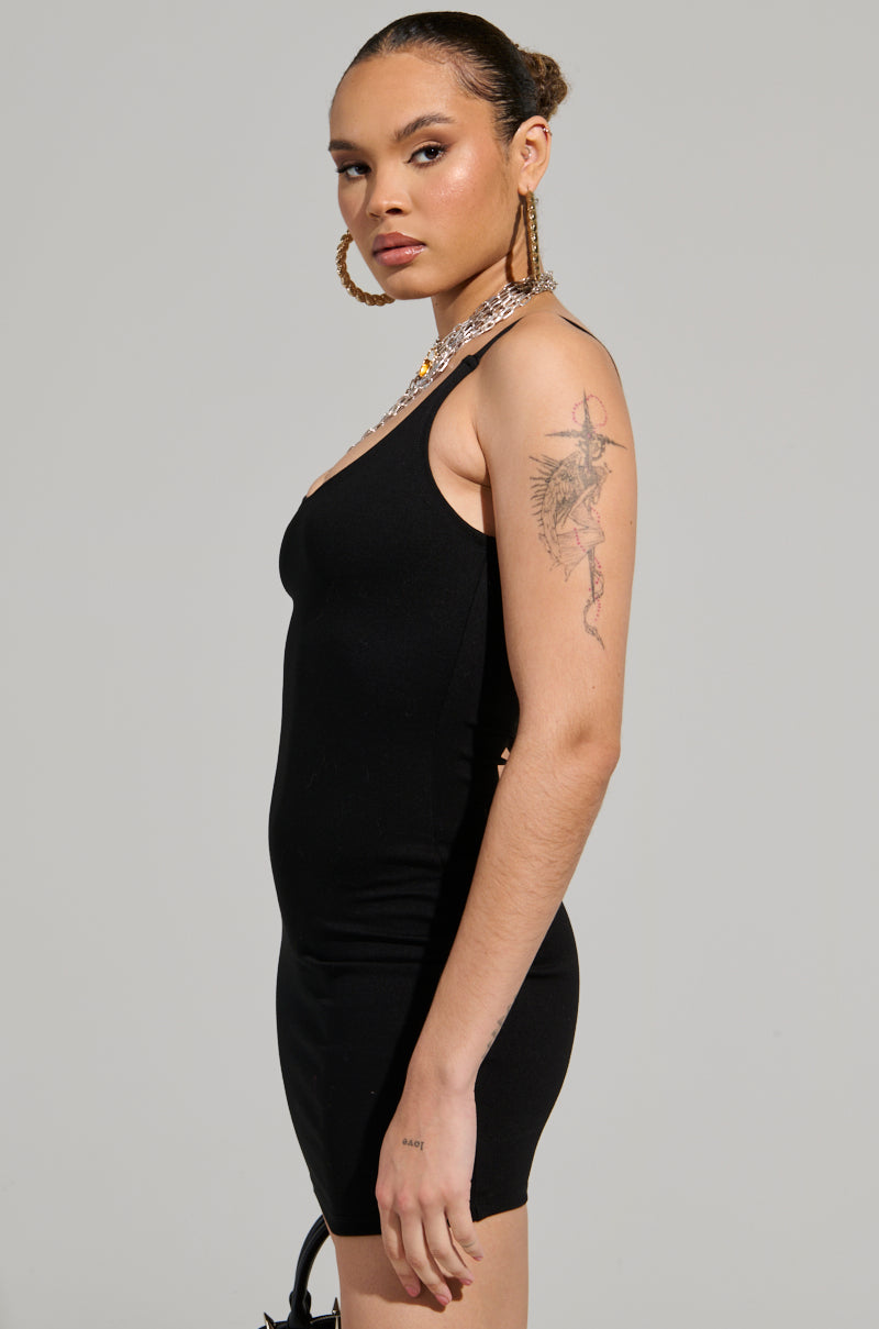 A person with braided hair and hoop earrings poses in the stylish REYLA Mini Bodycon Dress in Black, revealing a tattoo on their upper arm. They stand against a plain gray background.