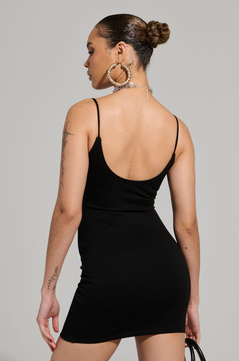 A person wearing the REYLA MINI BODYCON DRESS IN BLACK stands with their back to the camera, adorned with large hoop earrings, a chain necklace, and tattoos on their left arm. Their hair is styled in a bun.