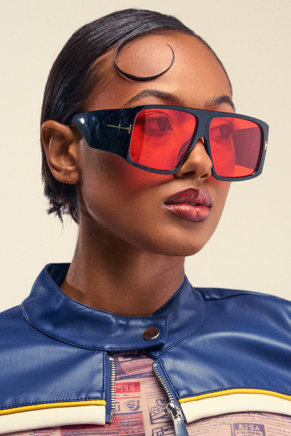 A woman with short, sleek hair wears the MIND YOUR BUSINESS SUNGLASSES—oversized and black with a retro style—paired with a blue jacket and a patterned shirt, gazing slightly to the side against a neutral background.