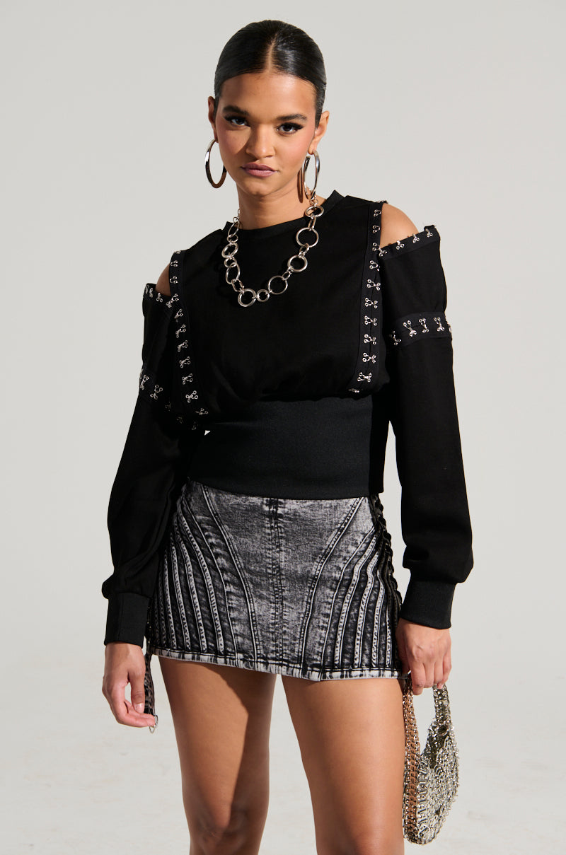 A person with slicked-back hair, large hoop earrings, and a serious expression is wearing an UNDER CONTROL SWEATSHIRT featuring cutout shoulders and faux silver metallic hardware. They also sport a short, metallic-patterned skirt and carry a small chain-link handbag.