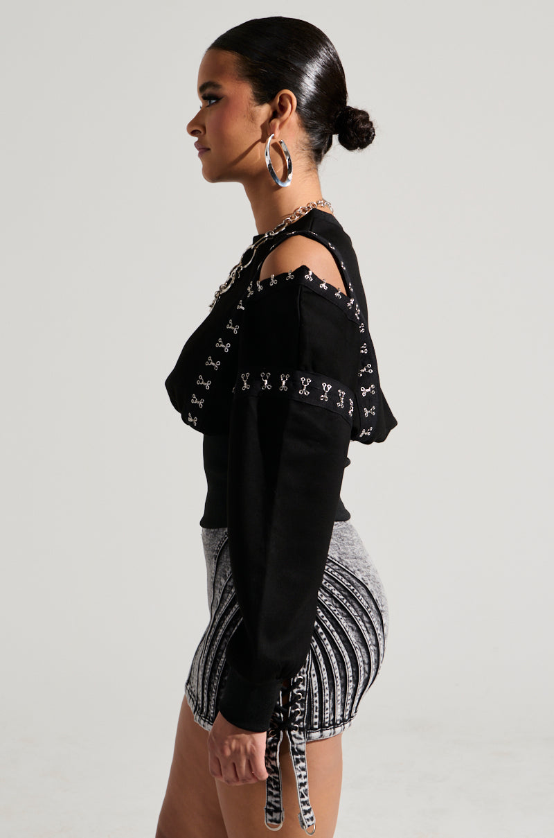 A woman in profile, sporting the UNDER CONTROL SWEATSHIRT featuring cut-out shoulders and faux silver metallic hardware, is paired with high-waisted, black and white striped shorts. Her hair is slicked back into a low bun, complemented by large hoop earrings. The background remains neutral.