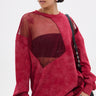 A woman in oversized sunglasses and statement earrings poses in the OPEN FOR BUSINESS SWEATSHIRT IN BURGUNDY with rhinestone mesh paneling over a black bra, styled with light-wash distressed jeans and a black shoulder bag.