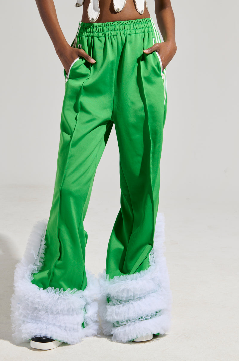 A person is casually standing with hands in pockets, sporting the MODERN JAM TRACK PANT WITH TULLE IN GREEN, which features bright green high-rise design, accented by white fur trim at the bottom and striking white stripes on the sides against a plain background.