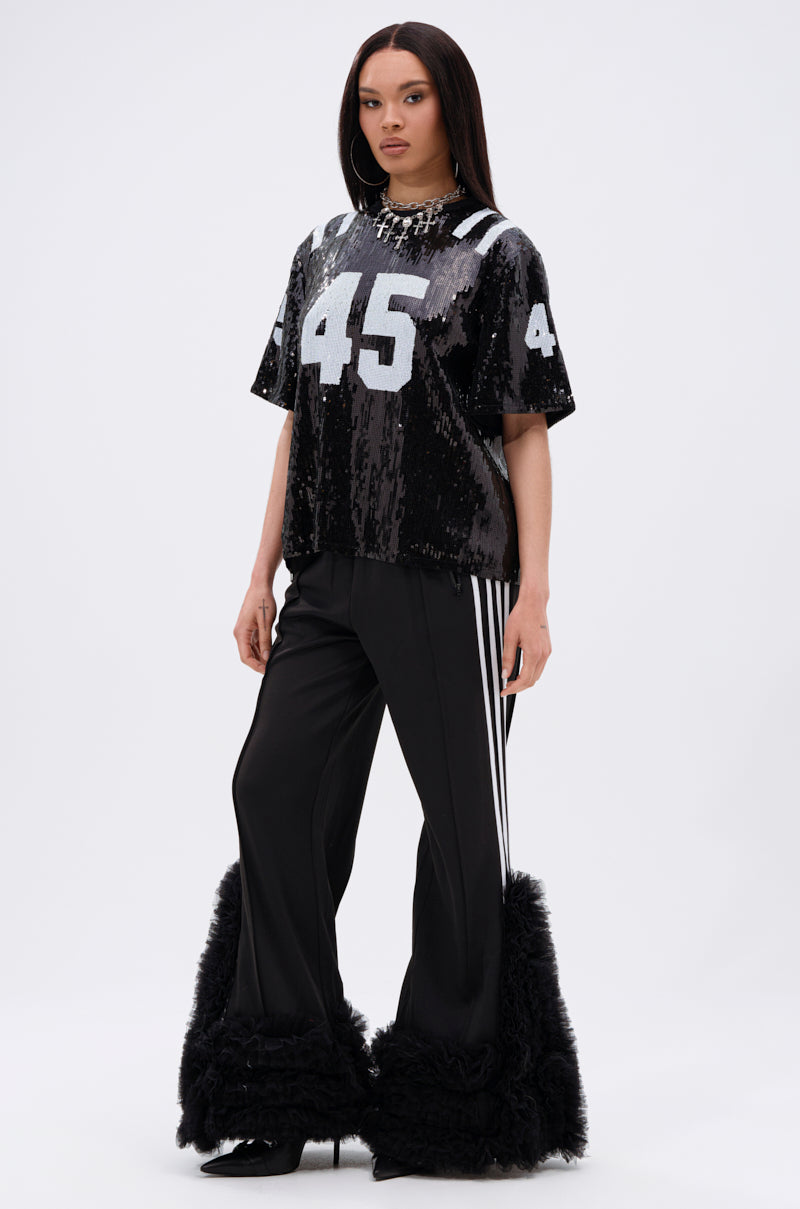 A woman faces forward in a black sequined AKIRA Label jersey, numbered 45 in white, and MODERN JAM TRACK PANT WITH TULLE IN BLACK, featuring white side stripes and fluffy tulle embellishments, against a plain light background.