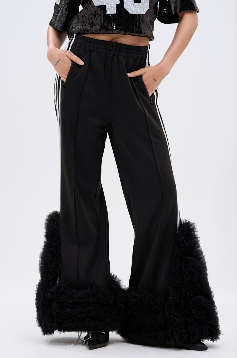 A person wears the MODERN JAM TRACK PANT WITH TULLE IN BLACK, featuring a wide-leg silhouette, white side stripes, and ruffled tulle cuffs, paired with a cropped black sequin top with white numbers. Hands are in pockets; face is not shown.