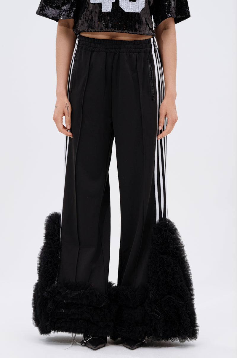 Wearing the MODERN JAM TRACK PANT WITH TULLE IN BLACK, which features black track pants with white side stripes and dramatic ruffled tulle on the lower legs. Only the wide-leg silhouette of the lower half is visible.