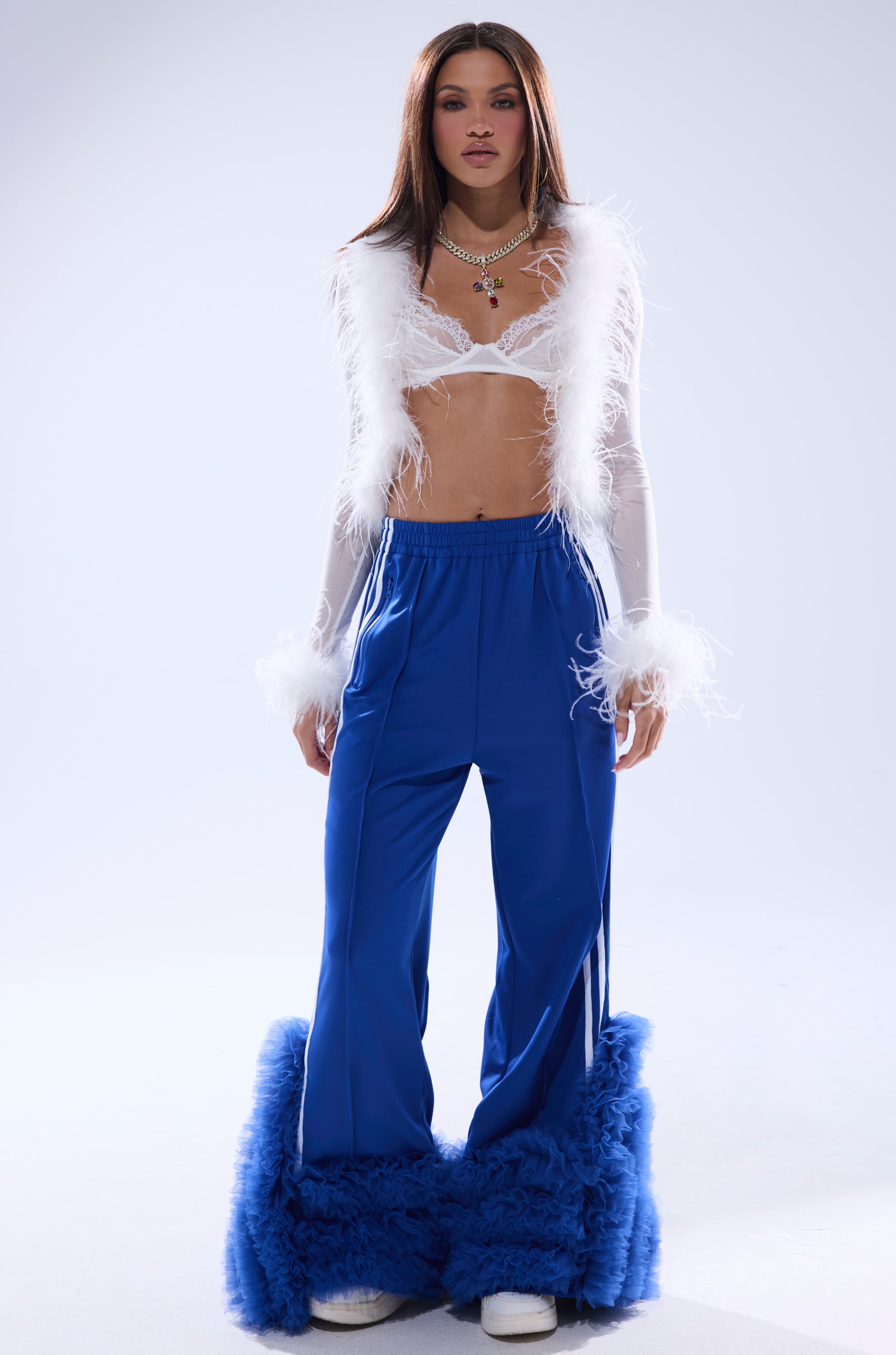 A woman faces forward against a plain light background, wearing a sheer white feathered crop top and the MODERN JAM TRACK PANT WITH TULLE IN ROYAL BLUE with fluffy hem trim, paired with white shoes and layered necklaces. 2