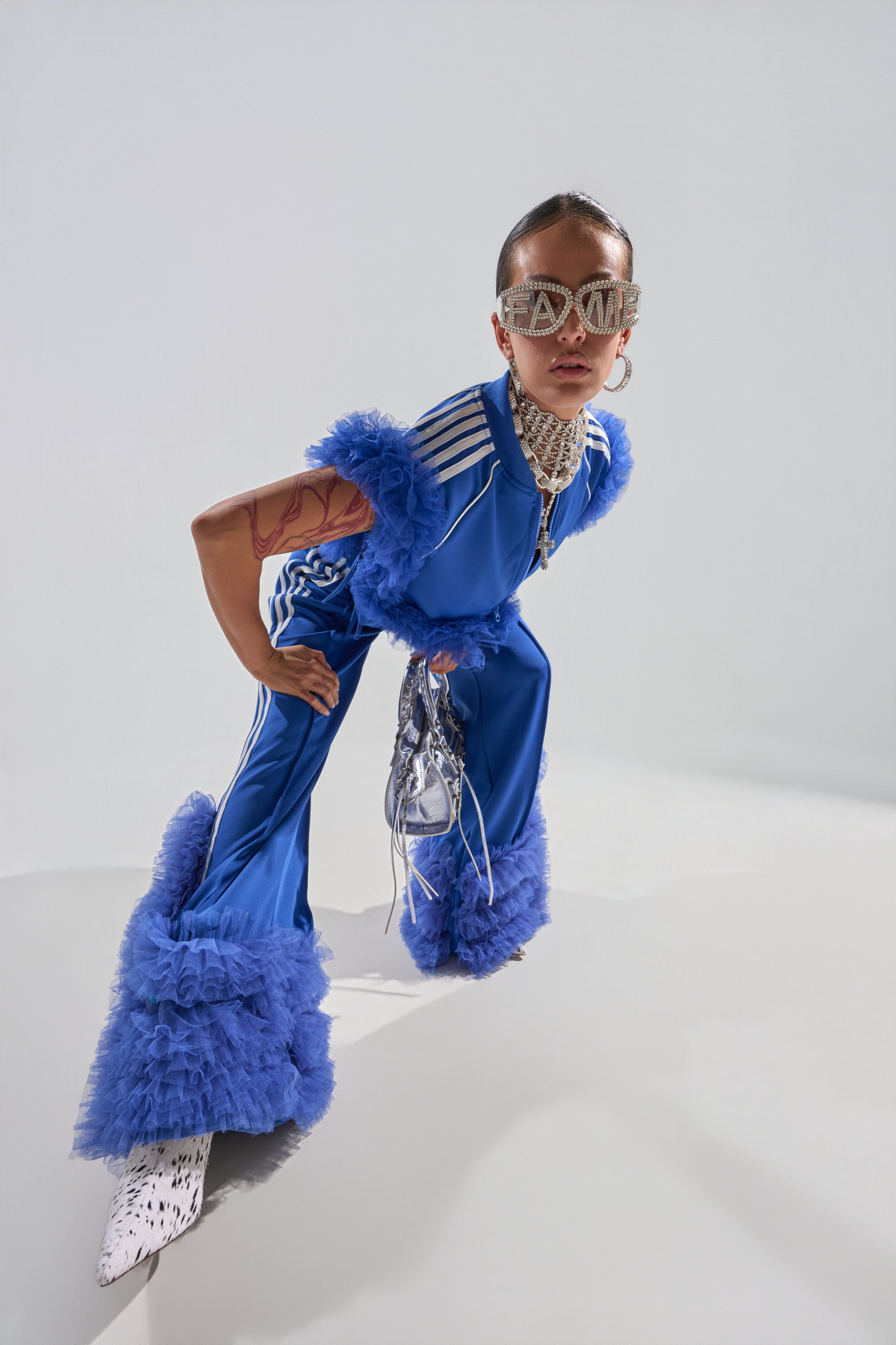 A person poses confidently in the MODERN JAM TRACK PANT WITH TULLE IN ROYAL BLUE, featuring ruffled tulle trim and white stripes. Silver accessories, oversized jeweled sunglasses, and patterned shoes complete this bold, avant-garde look. 0