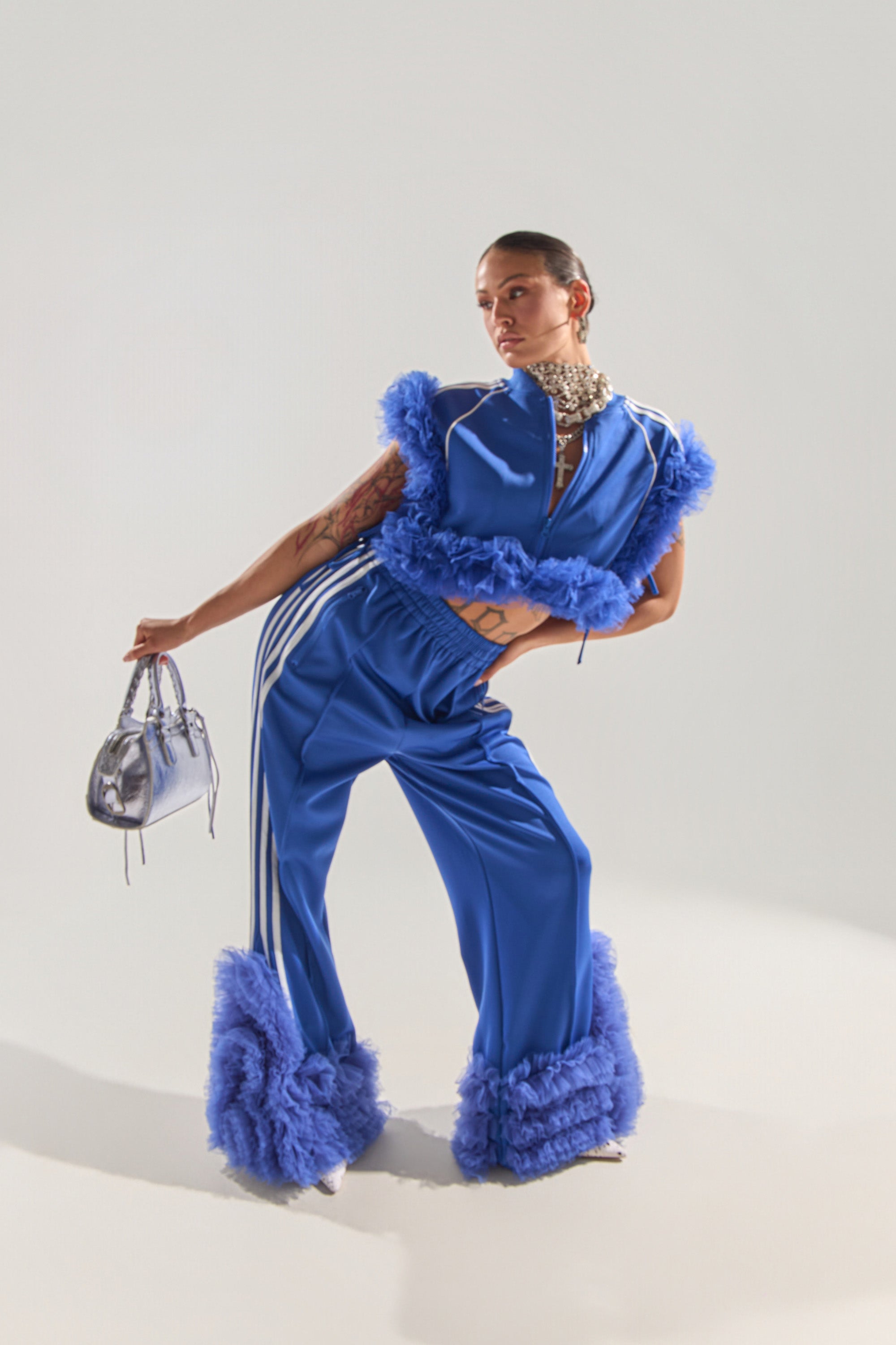 A person poses in the MODERN JAM TRACK PANT WITH TULLE IN ROYAL BLUE—featuring white stripes, flared bell bottoms, and fluffy trim—styled with silver heels, a metallic handbag, and a statement necklace against a plain light background. 1