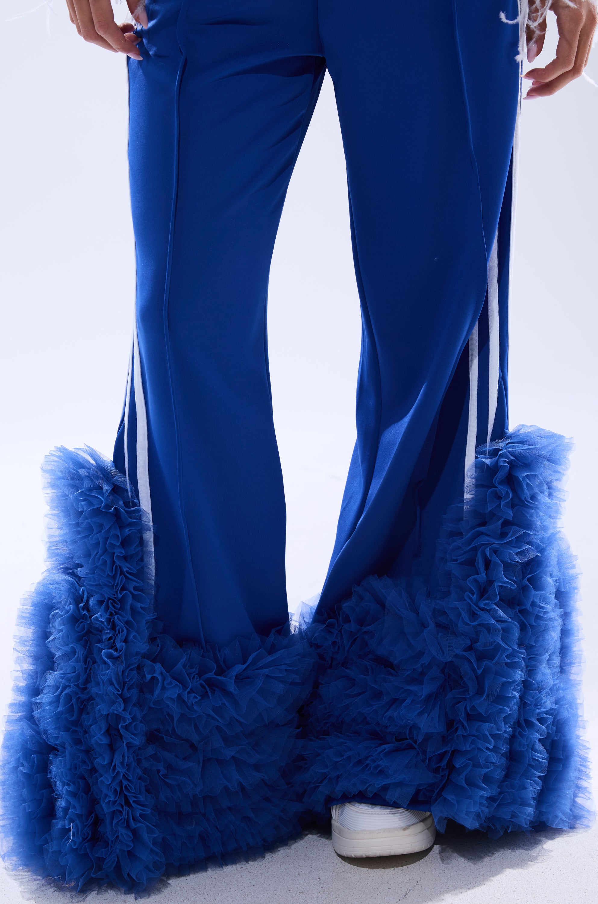 A person wears the MODERN JAM TRACK PANT WITH TULLE IN ROYAL BLUE, featuring dramatic ruffled tulle bell-bottom flares. Cropped above the waist, the image shows white sneakers partially visible on a light surface. 9