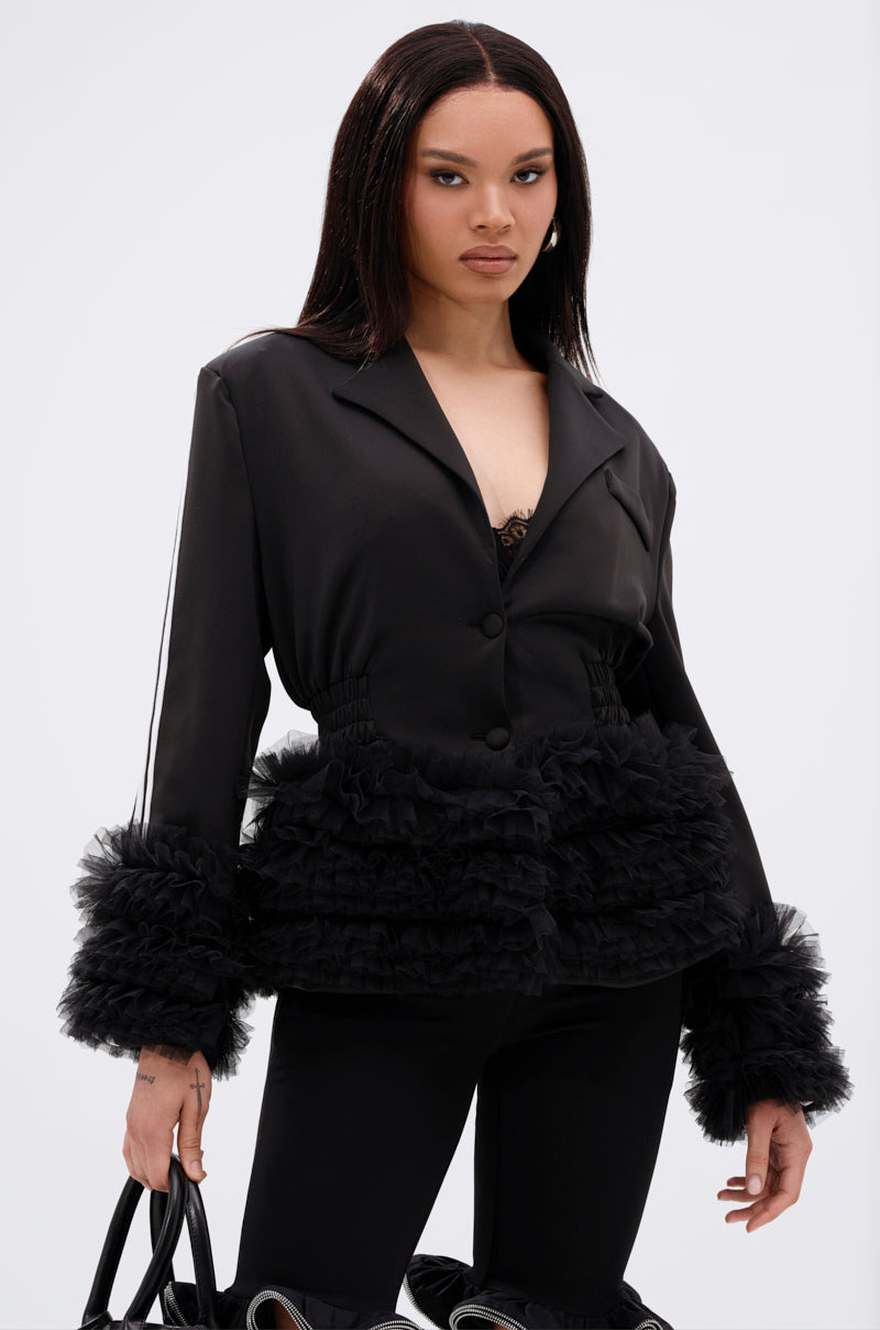 A woman with straight dark hair wears the BIG HIT TULLE TRIM BLAZER featuring ruffled tulle at the cuffs and waist, paired with black pants and a black handbag. She stands facing forward against a plain white background.