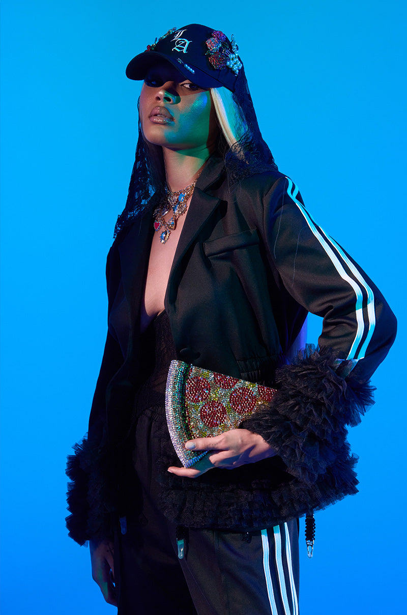 Against a blue background, a stylish person wears the BIG HIT TULLE TRIM BLAZER with furry cuffs and striped sleeves, accessorized with layered necklaces, a veiled cap, and a sparkling clutch bag.