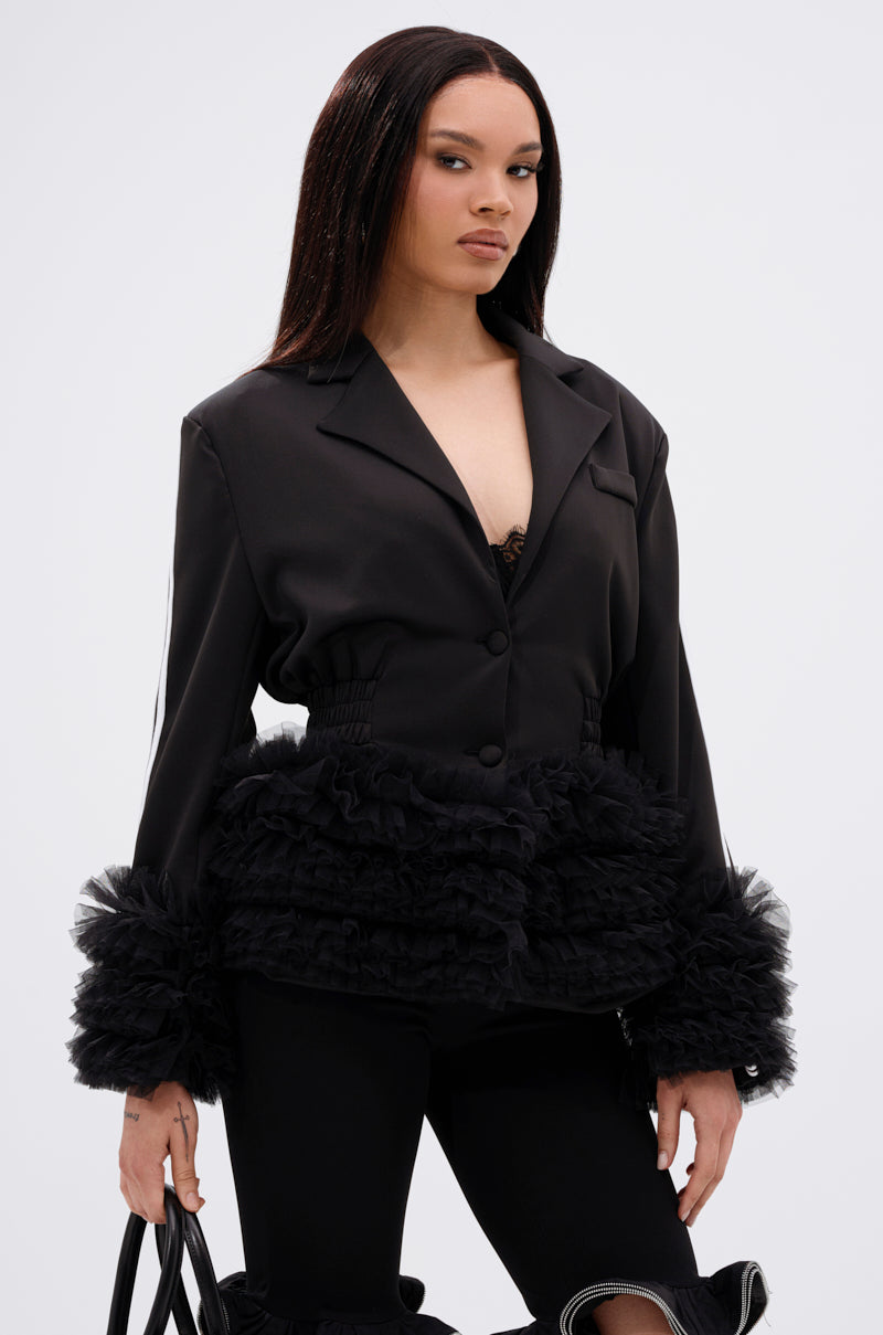 A woman with straight dark hair wears the BIG HIT TULLE TRIM BLAZER, featuring ruffled tulle at the waist and cuffs. She pairs it with black pants, holds a black bag, and stands against a plain white background.