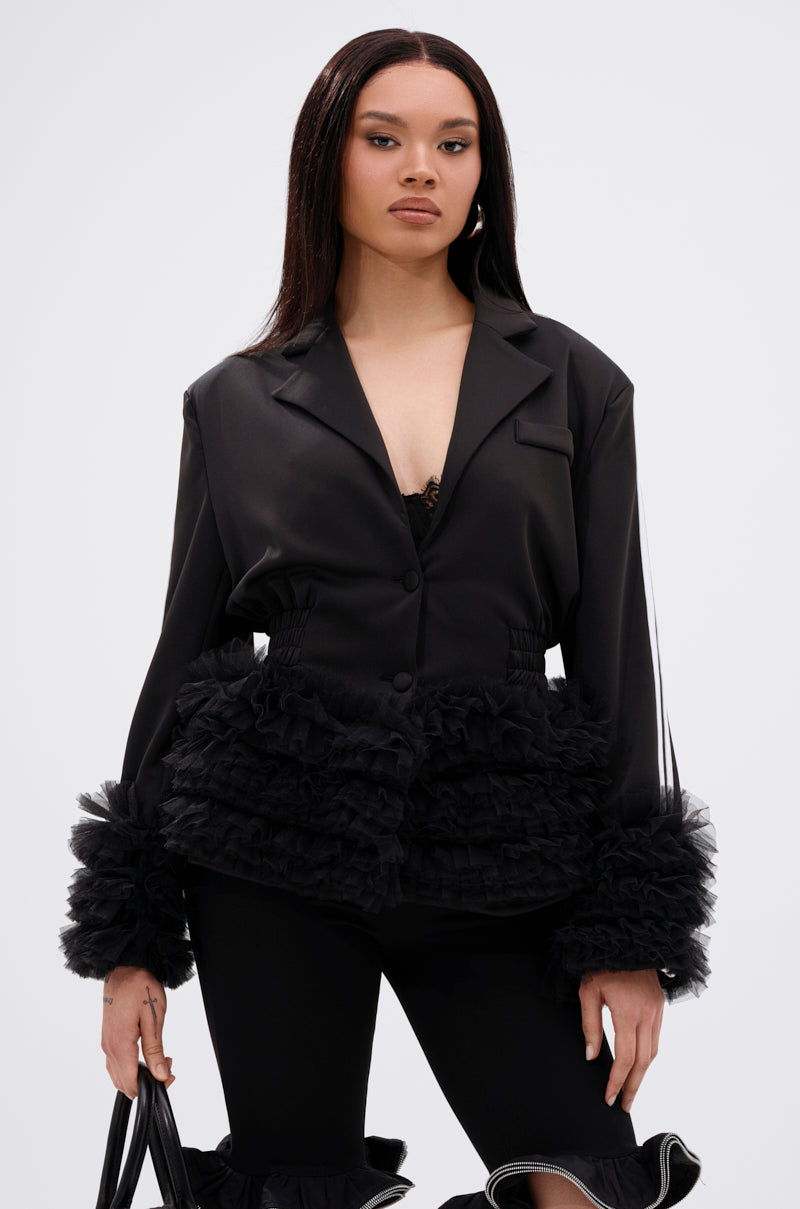 A woman with long straight hair and a neutral expression stands confidently in the BIG HIT TULLE TRIM BLAZER, featuring ruffled tulle details at the waist and sleeves, matching ruffle-accented pants, and holding a black bag.
