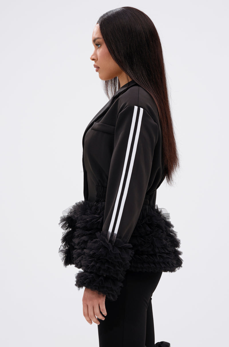 A woman in profile wears the BIG HIT TULLE TRIM BLAZER, featuring two white sleeve stripes and black ruffled tulle at the waist and cuffs. She has long, straight hair and stands against a plain white background.