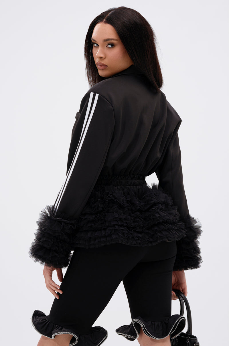 A woman in the BIG HIT TULLE TRIM BLAZER, featuring white sleeve stripes and ruffled cuffs, pairs it with matching black pants with ruffled hems. Holding a black handbag, she looks over her shoulder with a serious expression.