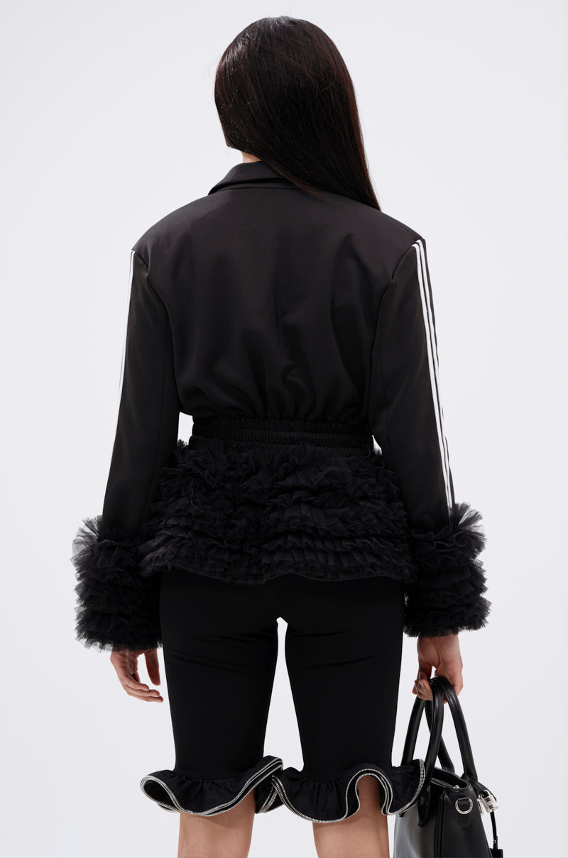 A person stands facing away, wearing the BIG HIT TULLE TRIM BLAZER with ruffled tulle at the waist and cuffs, paired with black knee-length shorts featuring ruffled hems. They hold a black handbag and have long, straight hair.