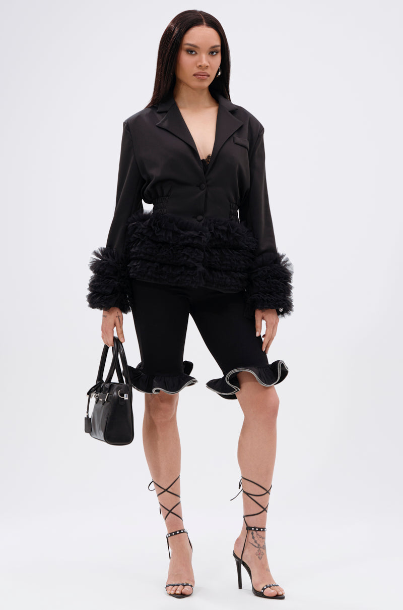 A woman with straight hair and a confident look poses against a plain background in the BIG HIT TULLE TRIM BLAZER, matching bermuda shorts, strappy heels, and a small black handbag.