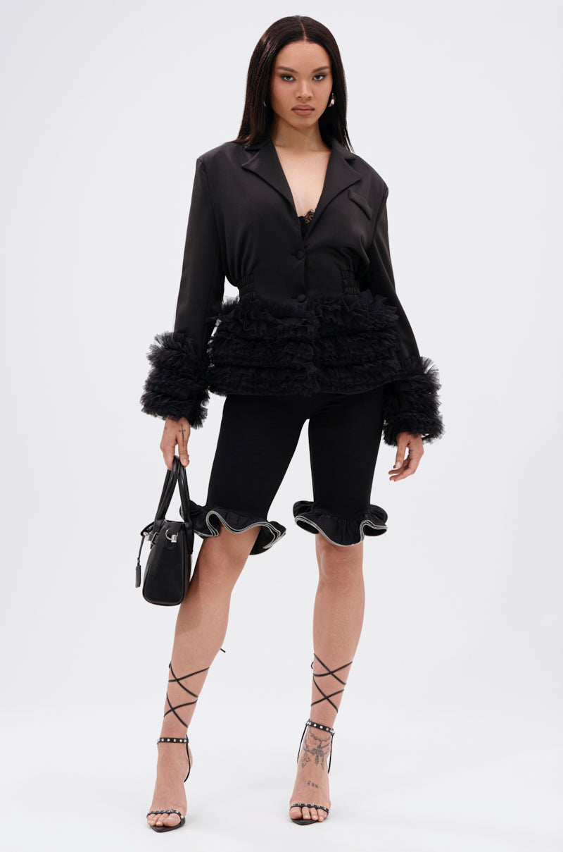 A woman stands confidently in the BIG HIT TULLE TRIM BLAZER with ruffled cuffs and tulle trim, paired with black cycling shorts, lace-up heels, and a black handbag against a plain white background.