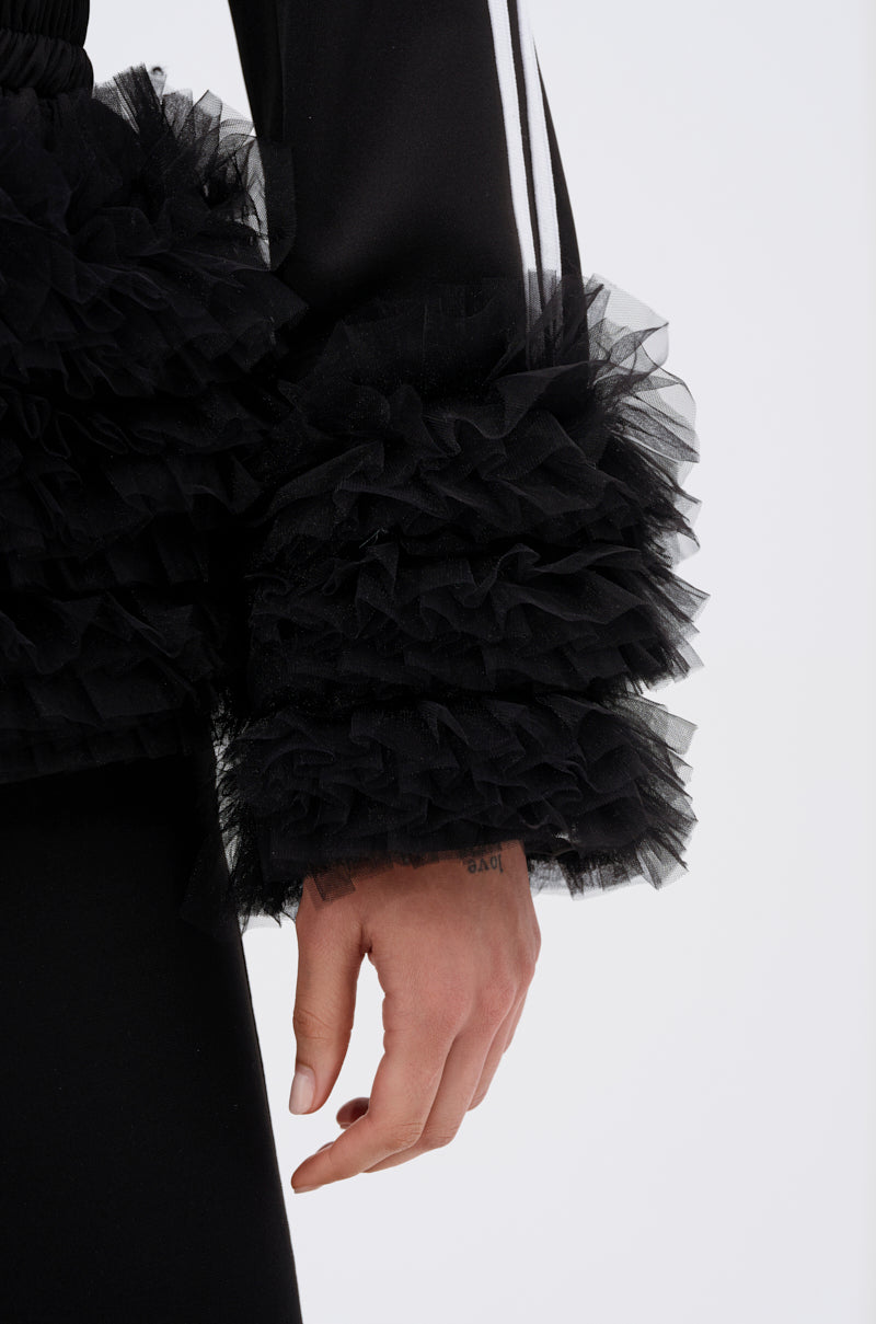 A close-up of a person’s arm wearing the BIG HIT TULLE TRIM BLAZER, featuring black streetwear style and dramatic layered tulle ruffle cuffs. Their hand is relaxed at their side against a plain white background.