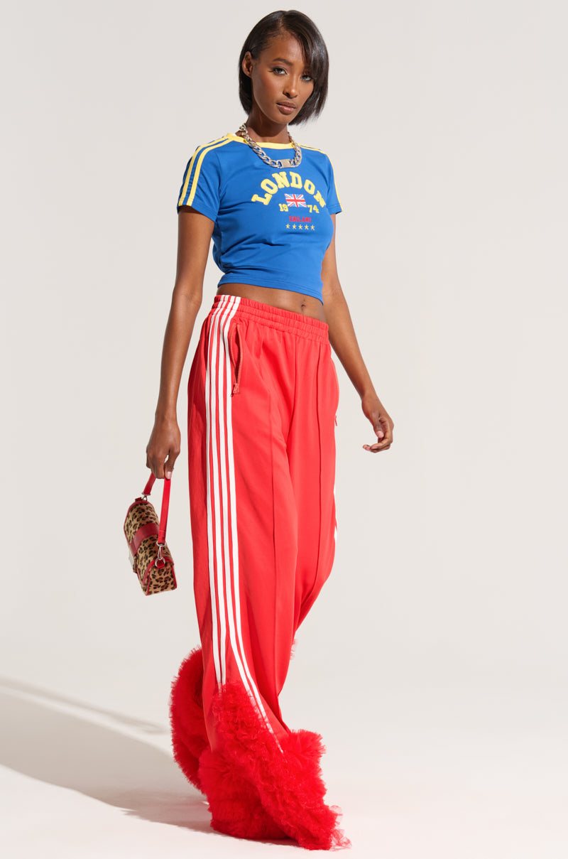 A fashionable person stands confidently wearing a blue "London" crop top, red Modern Jam Track Pant with Tulle featuring white stripes and a high-rise design, and red fluffy shoes. They hold a small leopard print handbag in their right hand. The background is plain white.